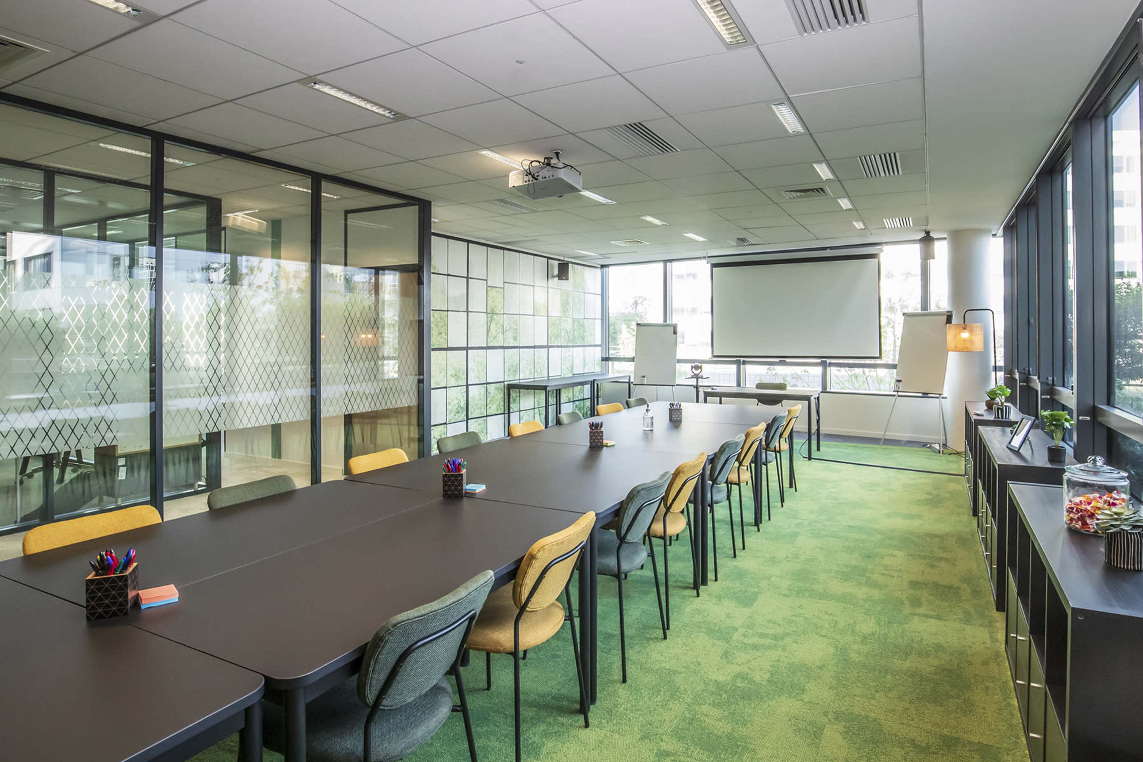 A modern conference room features a long table with chairs, large windows providing natural light, and a projector setup against a wall.