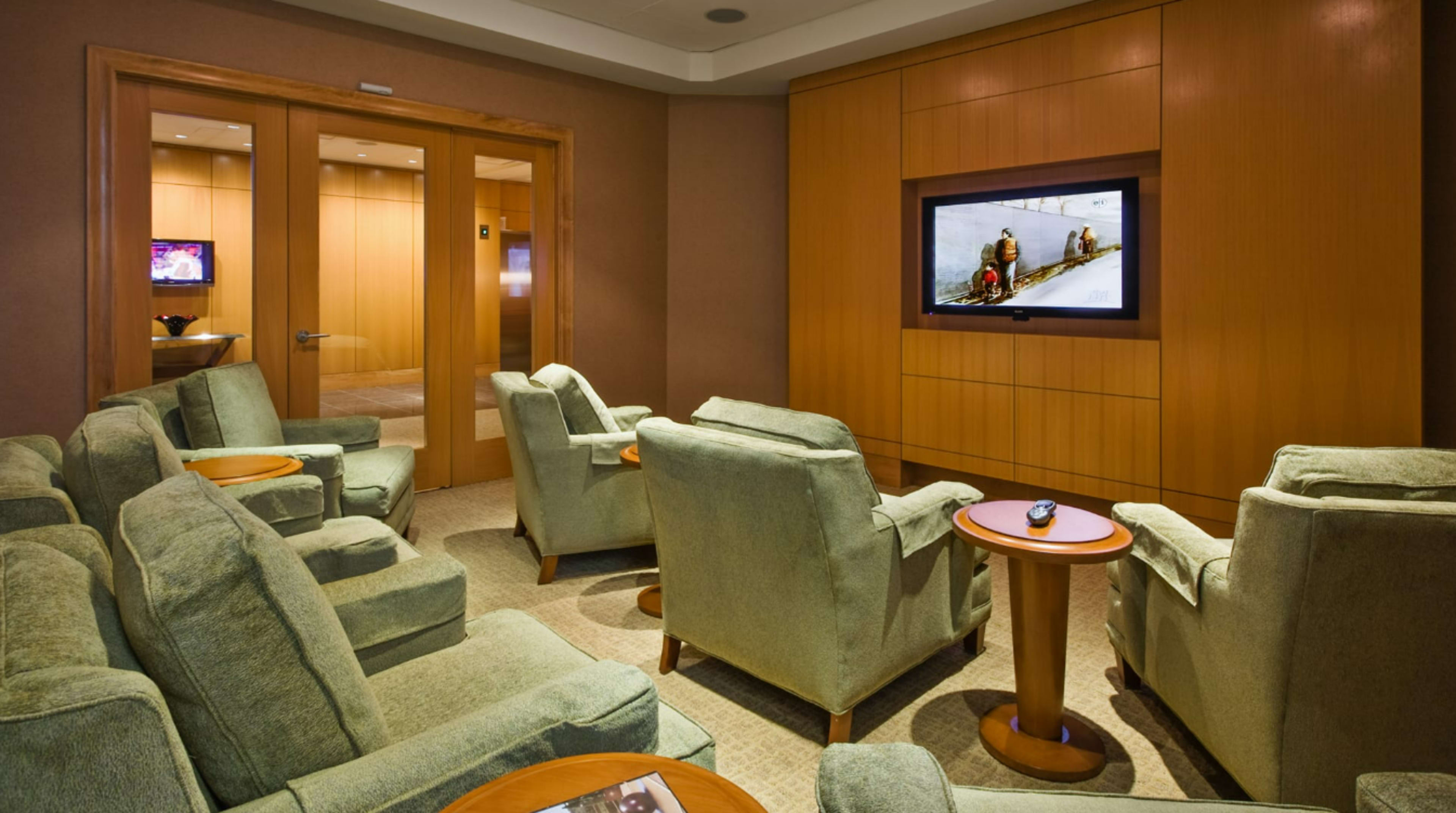 The image shows a lounge area with several green chairs arranged around a small table, facing a flat-screen TV mounted on the wall.