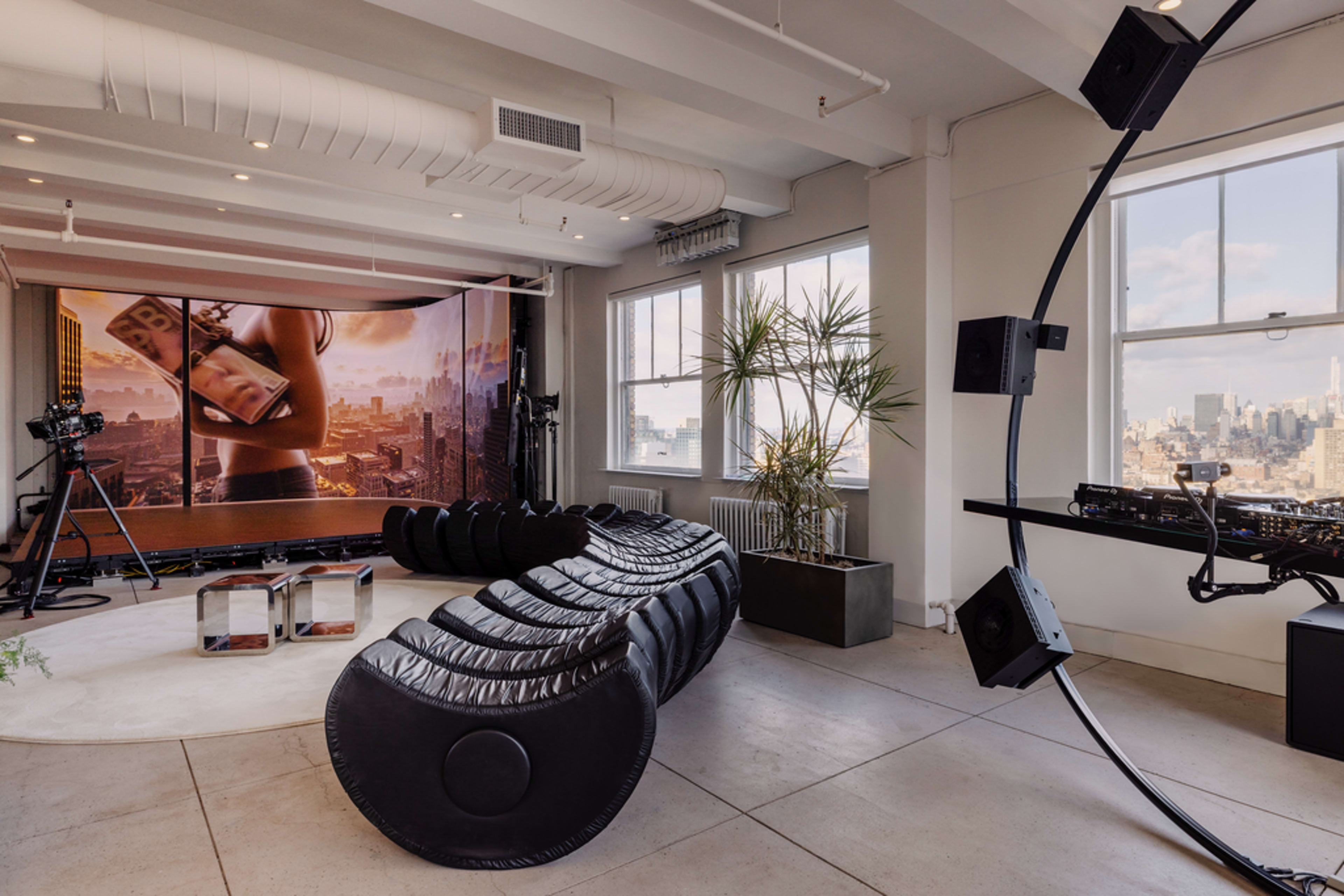 The image shows a modern lounge area with a curved black sofa, a large screen displaying a cityscape, and windows overlooking an urban skyline.