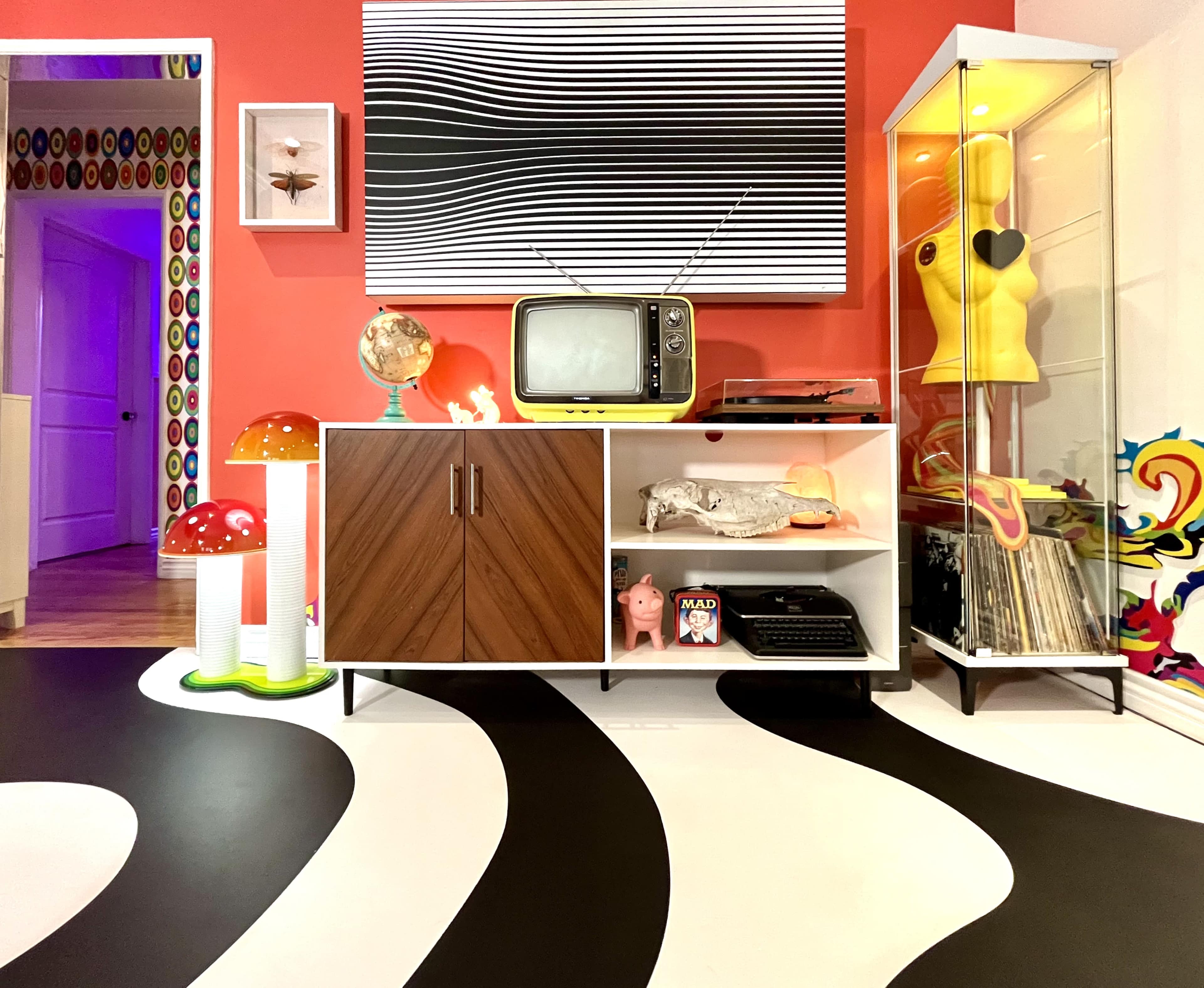 The scene features a colorful retro living room with a vintage television, a wooden cabinet, and decorative items like oversized mushrooms and a glass display case containing a yellow figurine.