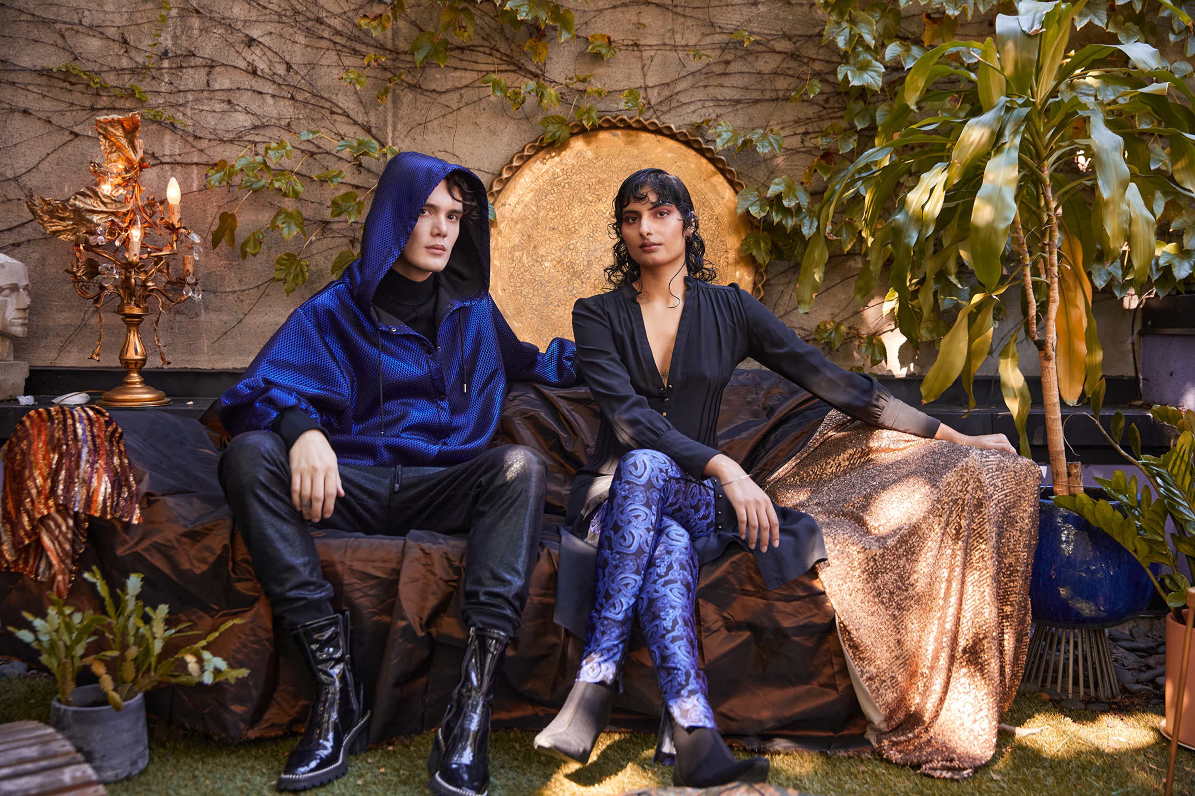 A man in a blue hoodie sits beside a woman in a black outfit, both posed on a couch surrounded by greenery and decorative elements.