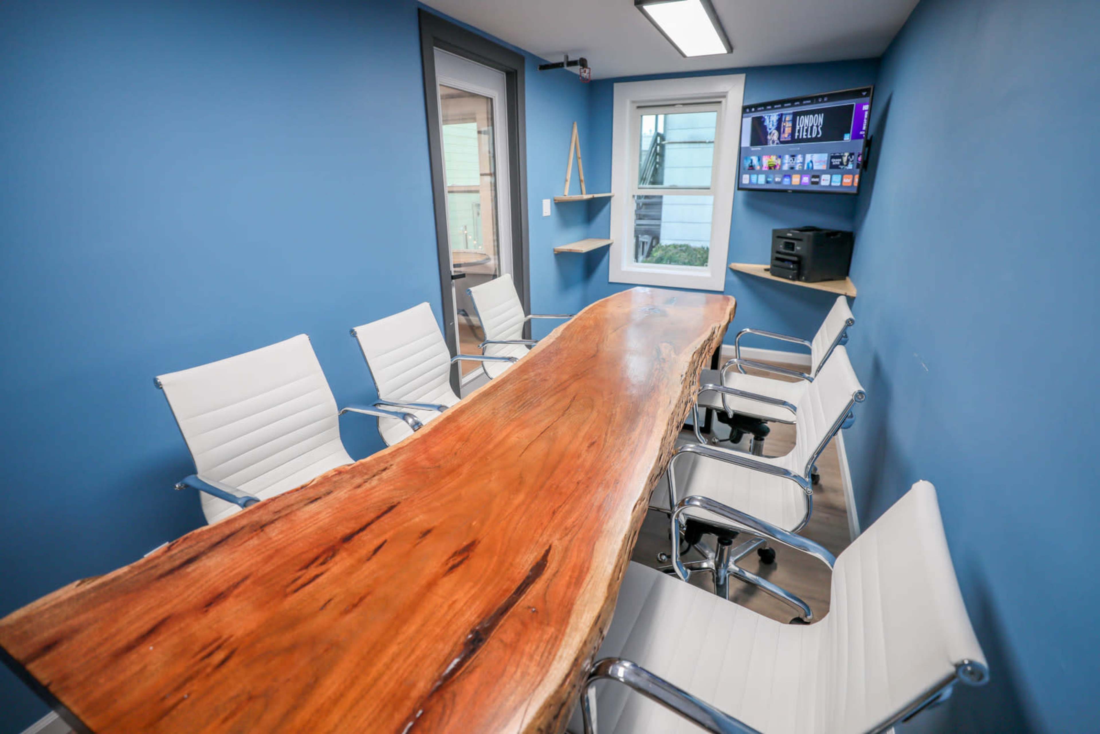 A modern conference room features a large wooden table surrounded by white chairs, with a blue wall and a window.