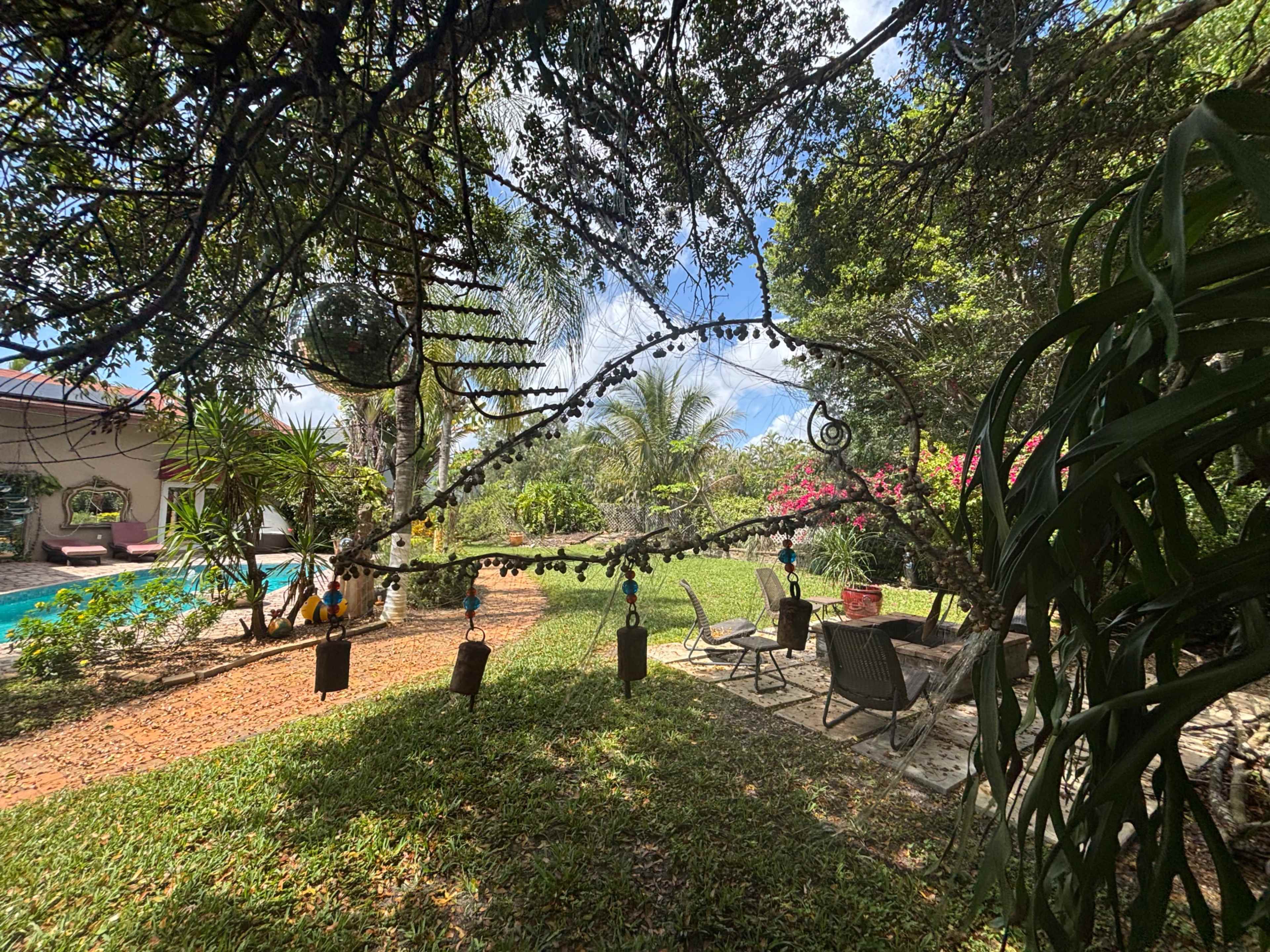 The scene shows a lush garden with a seating area surrounded by vibrant greenery and colorful flowers, next to a swimming pool.