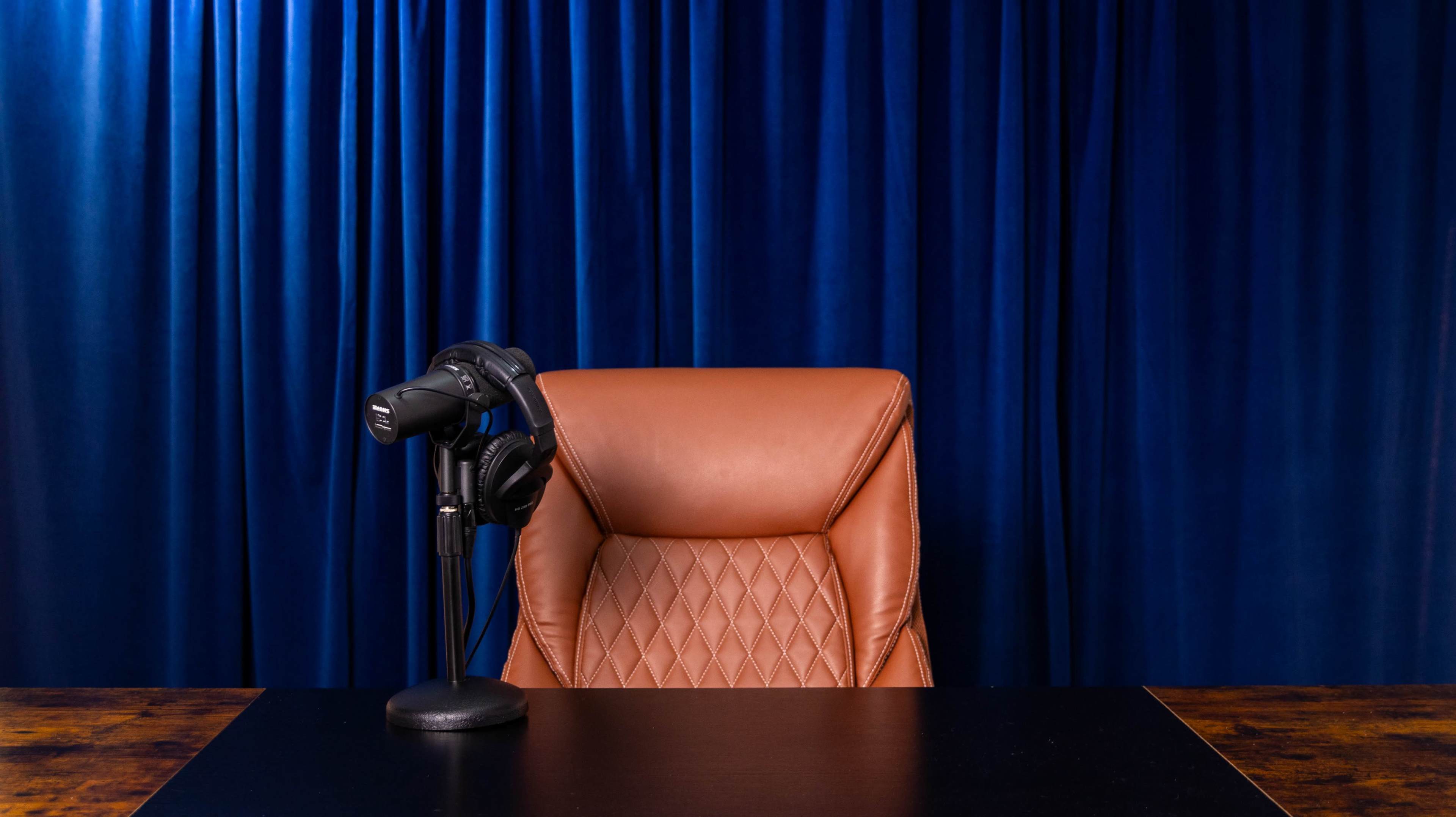 The image shows a brown leather chair and a microphone with headphones on a black desk, set against a blue curtain backdrop.
