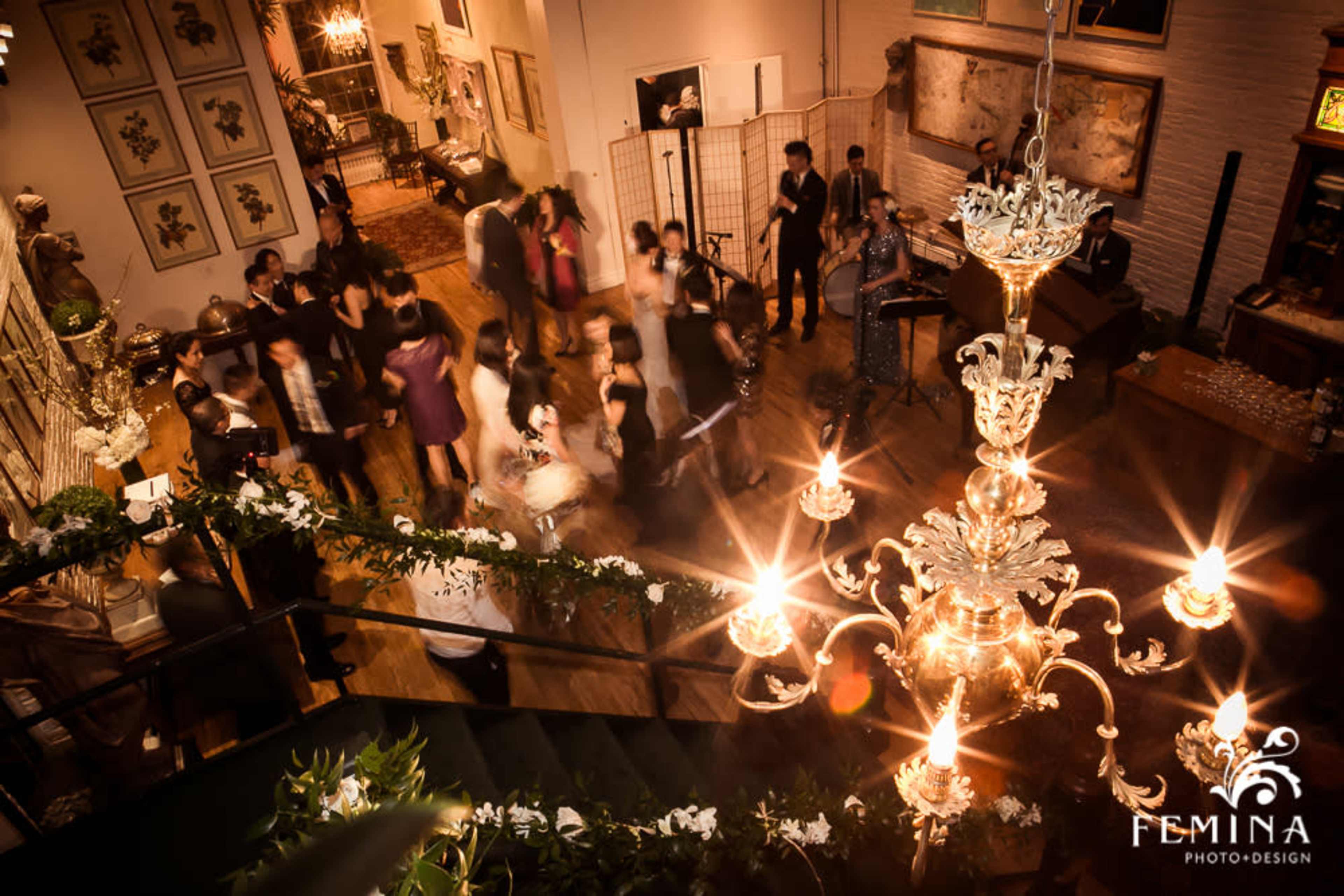 The image shows a lively indoor event with guests mingling and dancing under a large chandelier.