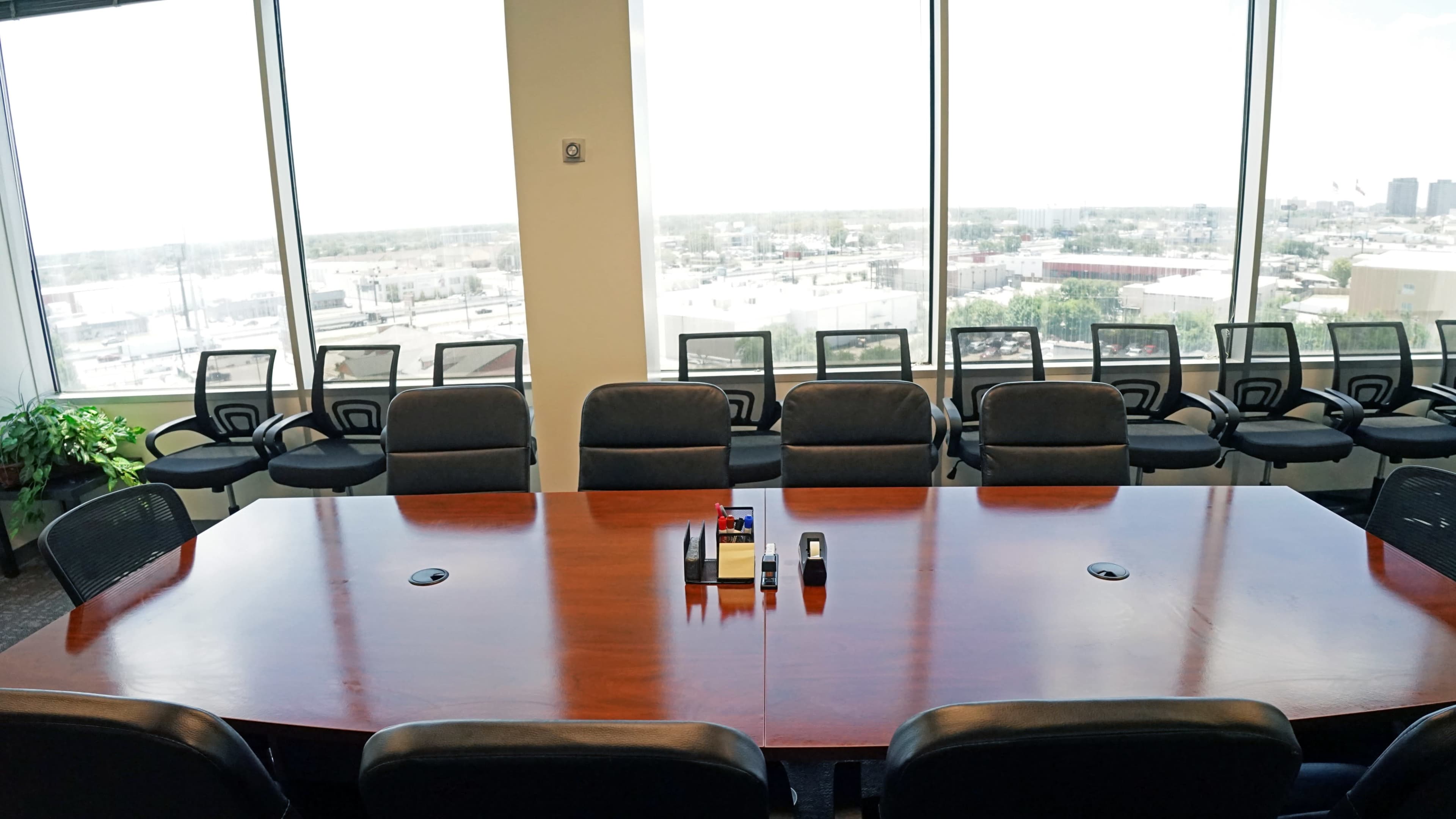 A large wooden conference table is set in a well-lit meeting room with panoramic windows overlooking an urban landscape and several office chairs arranged around it.