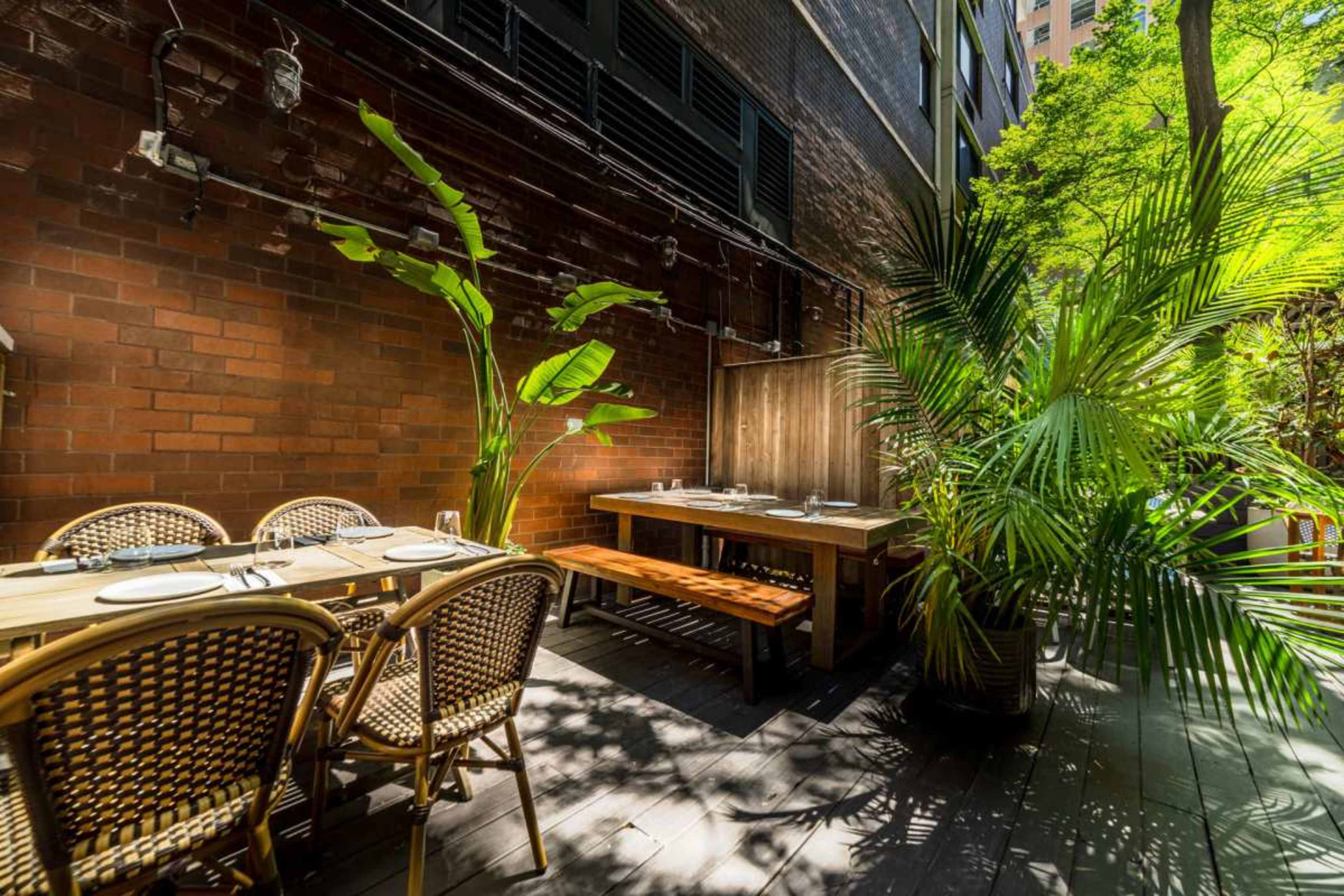 The image shows an outdoor dining area featuring wooden tables, chairs, and lush green plants against a brick wall.