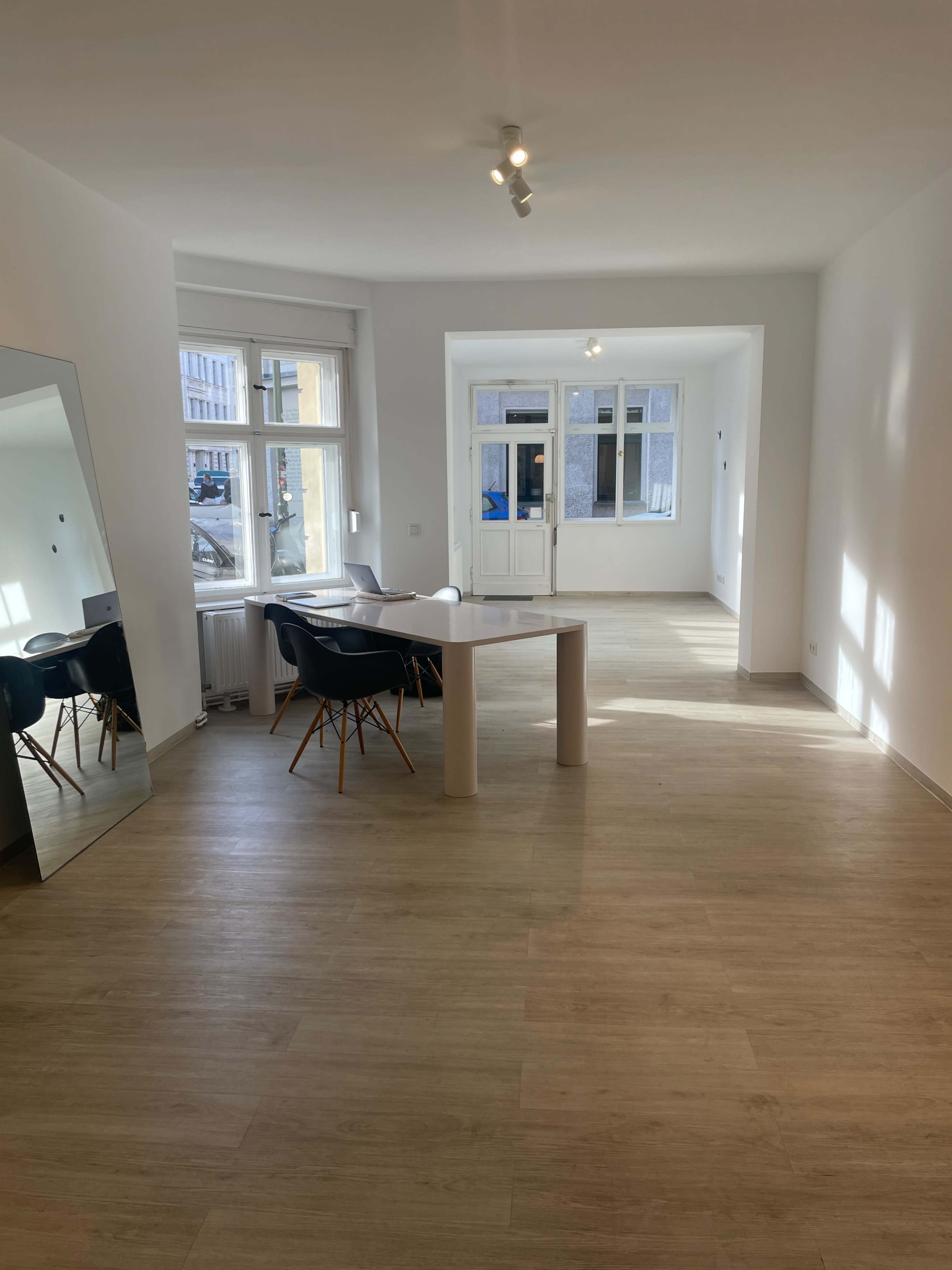The image shows a spacious, empty room with light-colored walls, a wooden floor, a table with chairs, and large windows allowing natural light to enter.