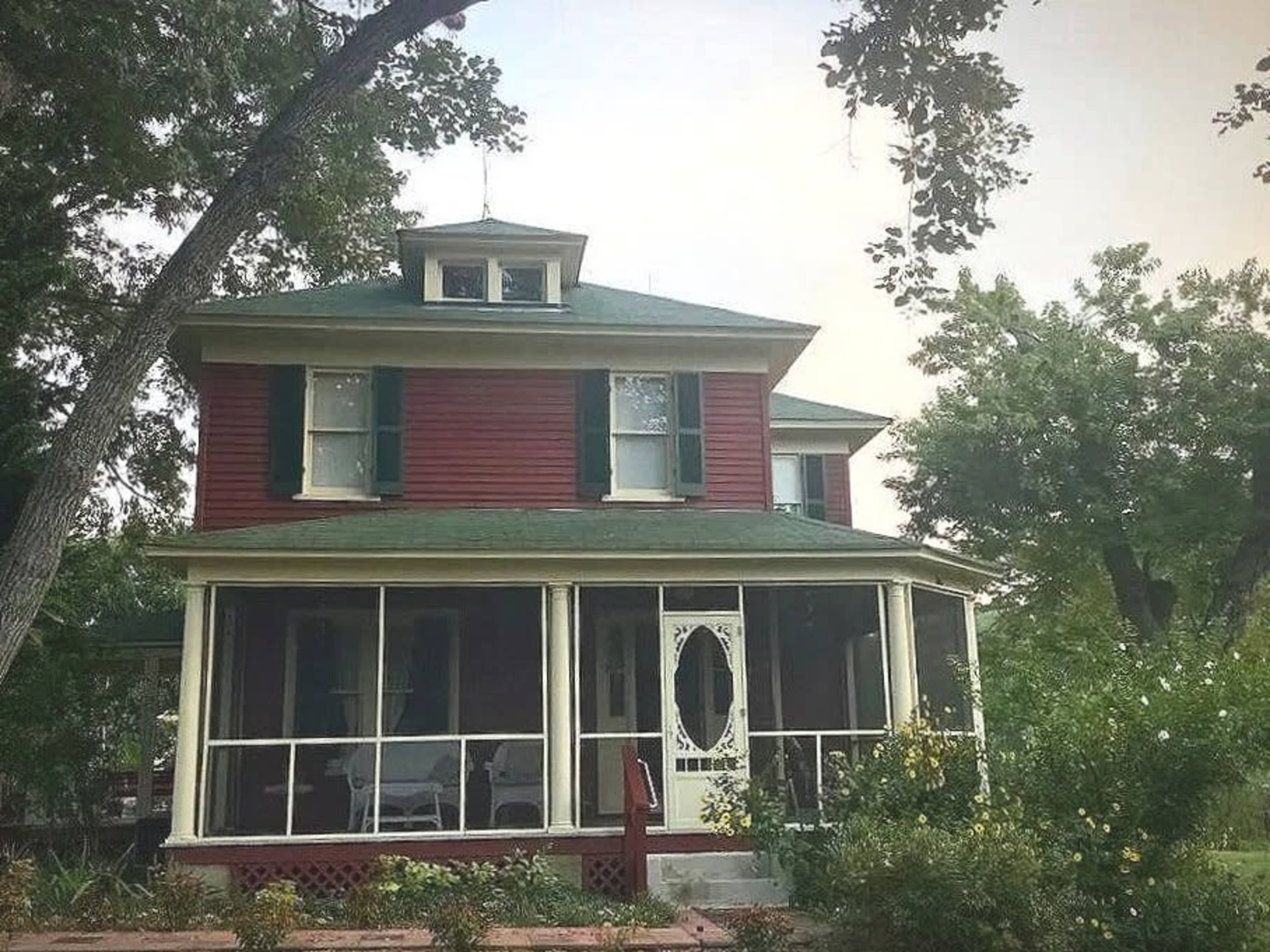 The image shows a red two-story house with green shutters and a covered porch surrounded by trees and greenery.
