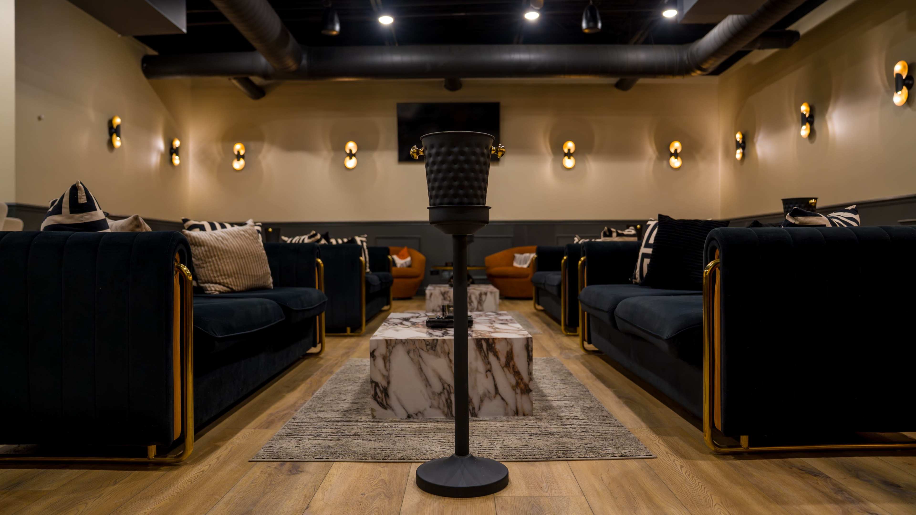 The image shows a stylish lounge area featuring dark velvet sofas, a marble coffee table, and warm lighting fixtures on the walls.