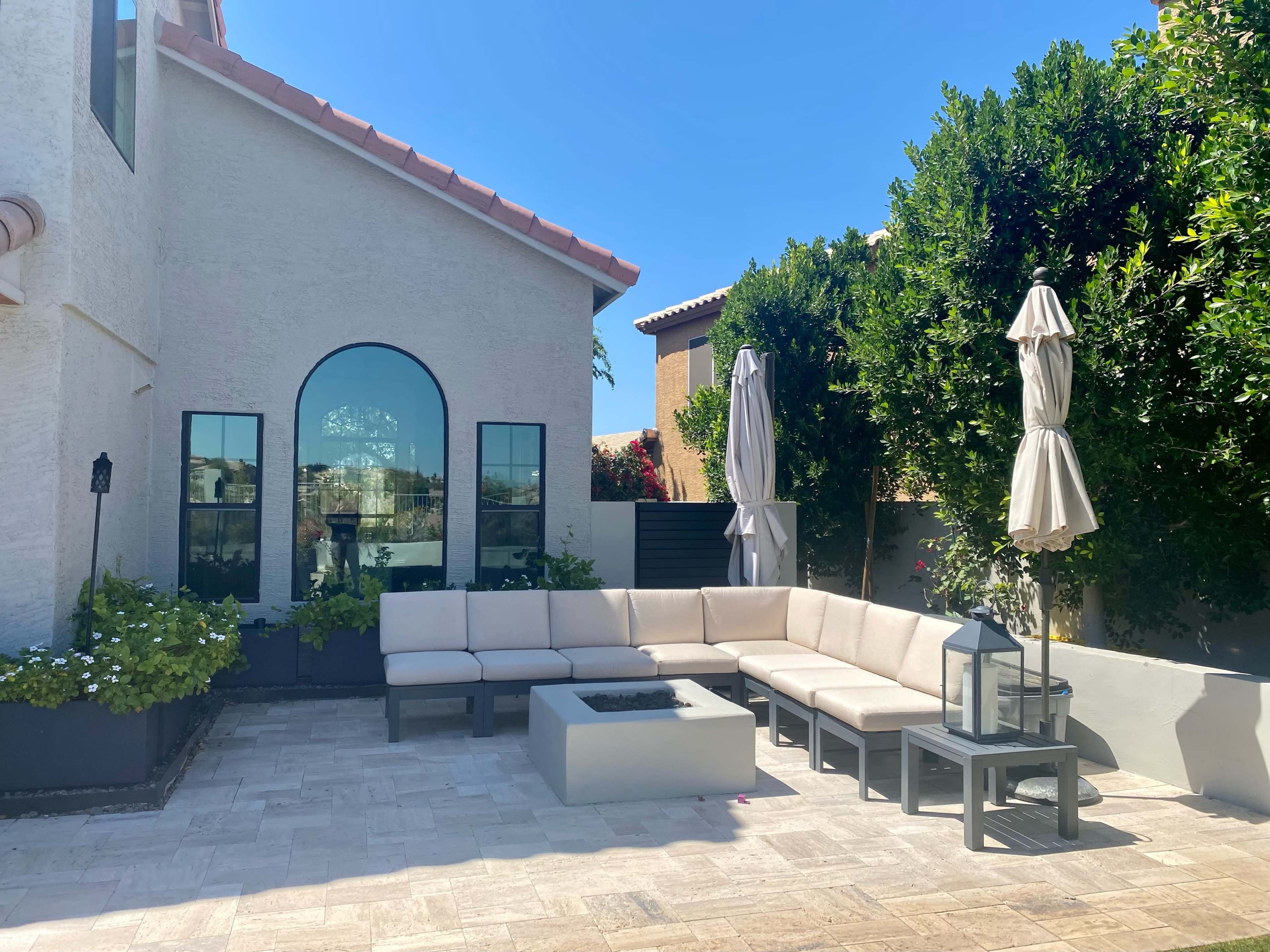 A shaded outdoor seating area with a modern sectional couch, a fire pit, and patio umbrellas surrounded by greenery.