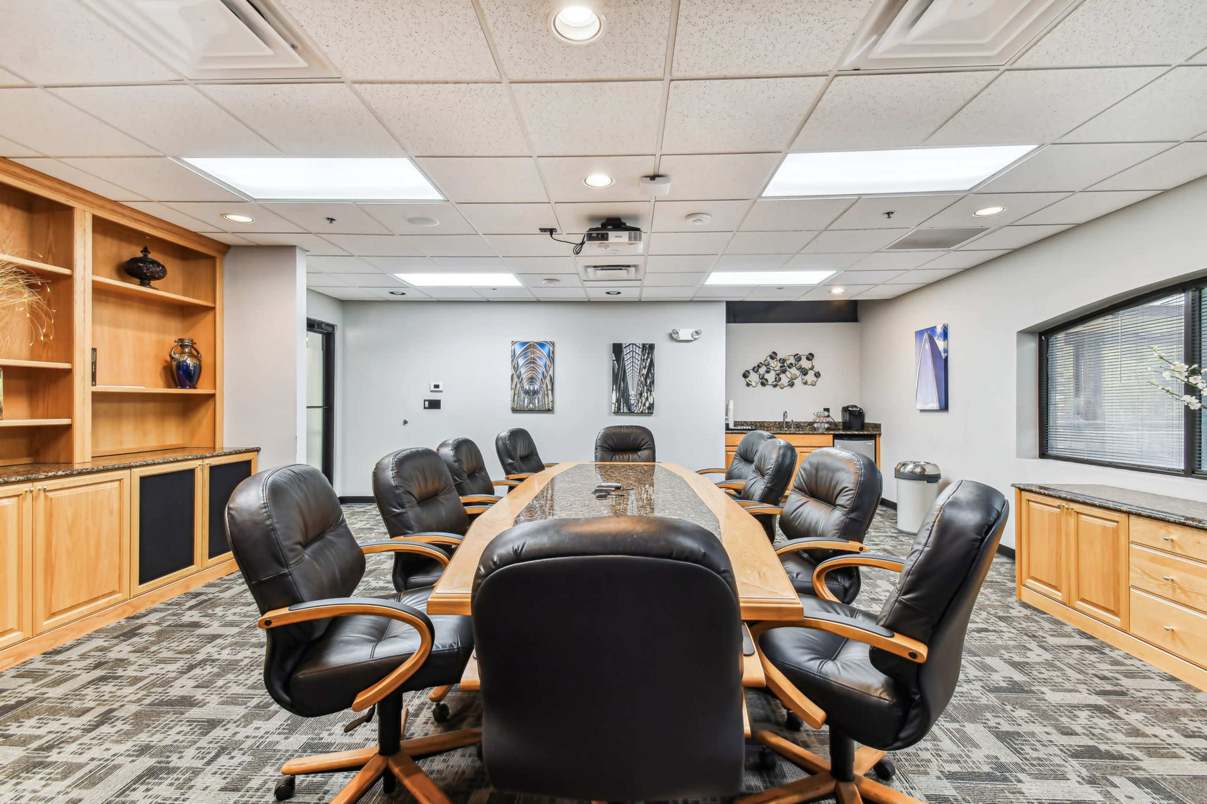 The image shows a spacious conference room featuring a long wooden table surrounded by black rolling chairs, with built-in cabinetry and modern lighting.