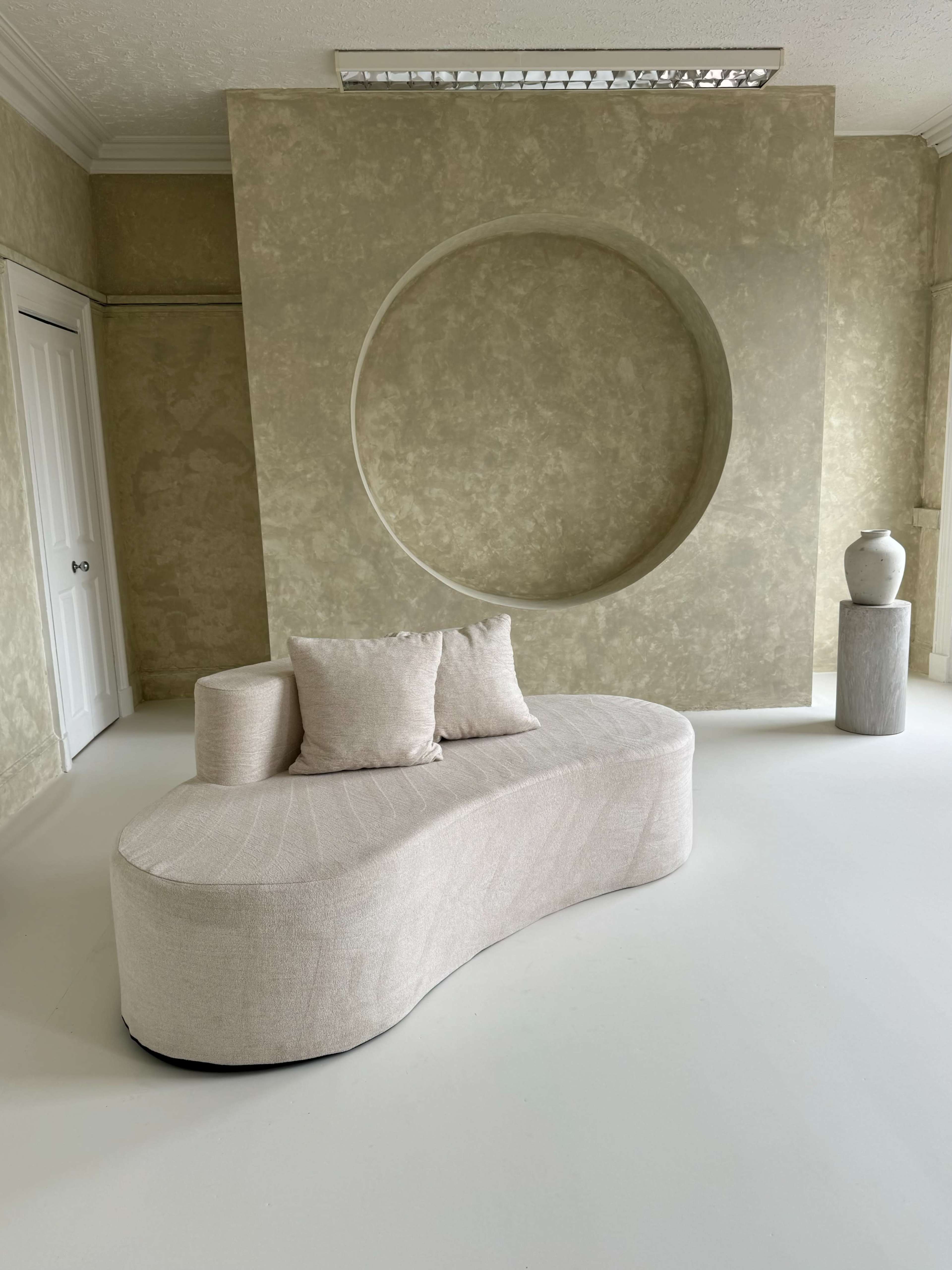 The image shows a minimalist interior with a curved beige sofa, a round wall niche, and a simple vase on a cylindrical pedestal.