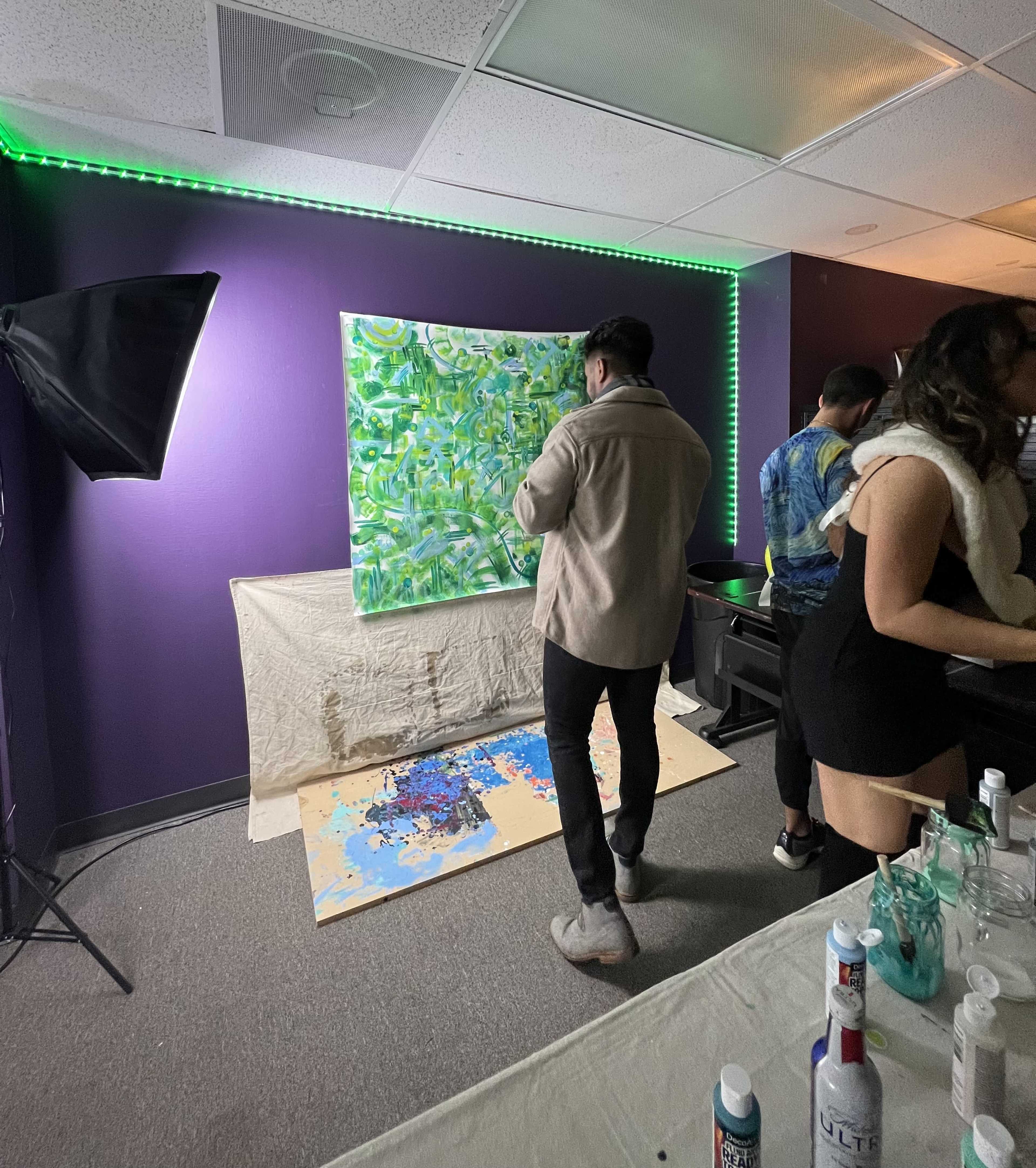 A person stands in front of a large painting on a purple wall, while others gather around a table with various bottles and art materials.