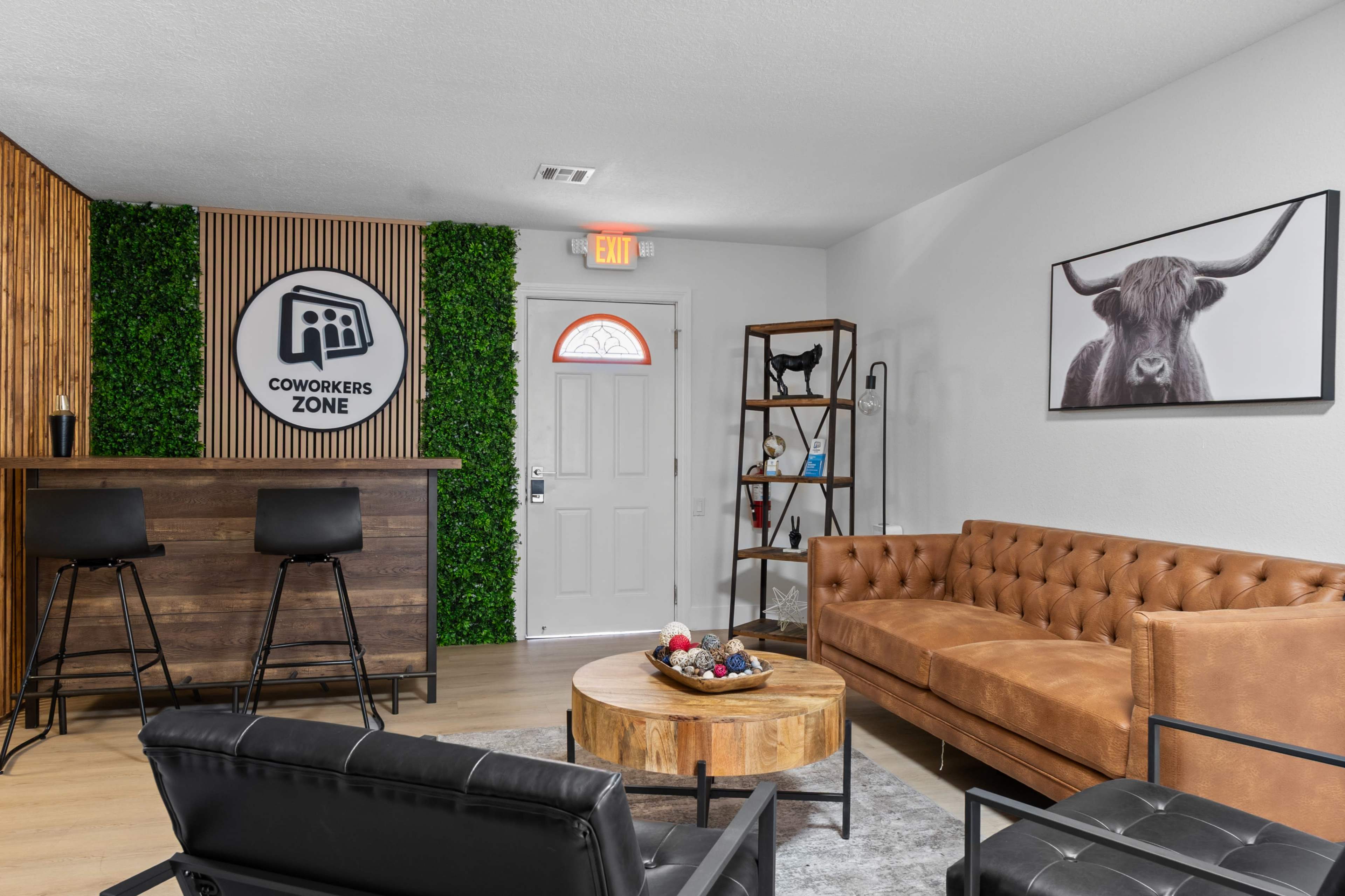 The image shows a modern coworking space featuring a brown leather sofa, a round wooden coffee table, black seating, and a bar area with a logo that reads "COWORKERS ZONE" surrounded by greenery.