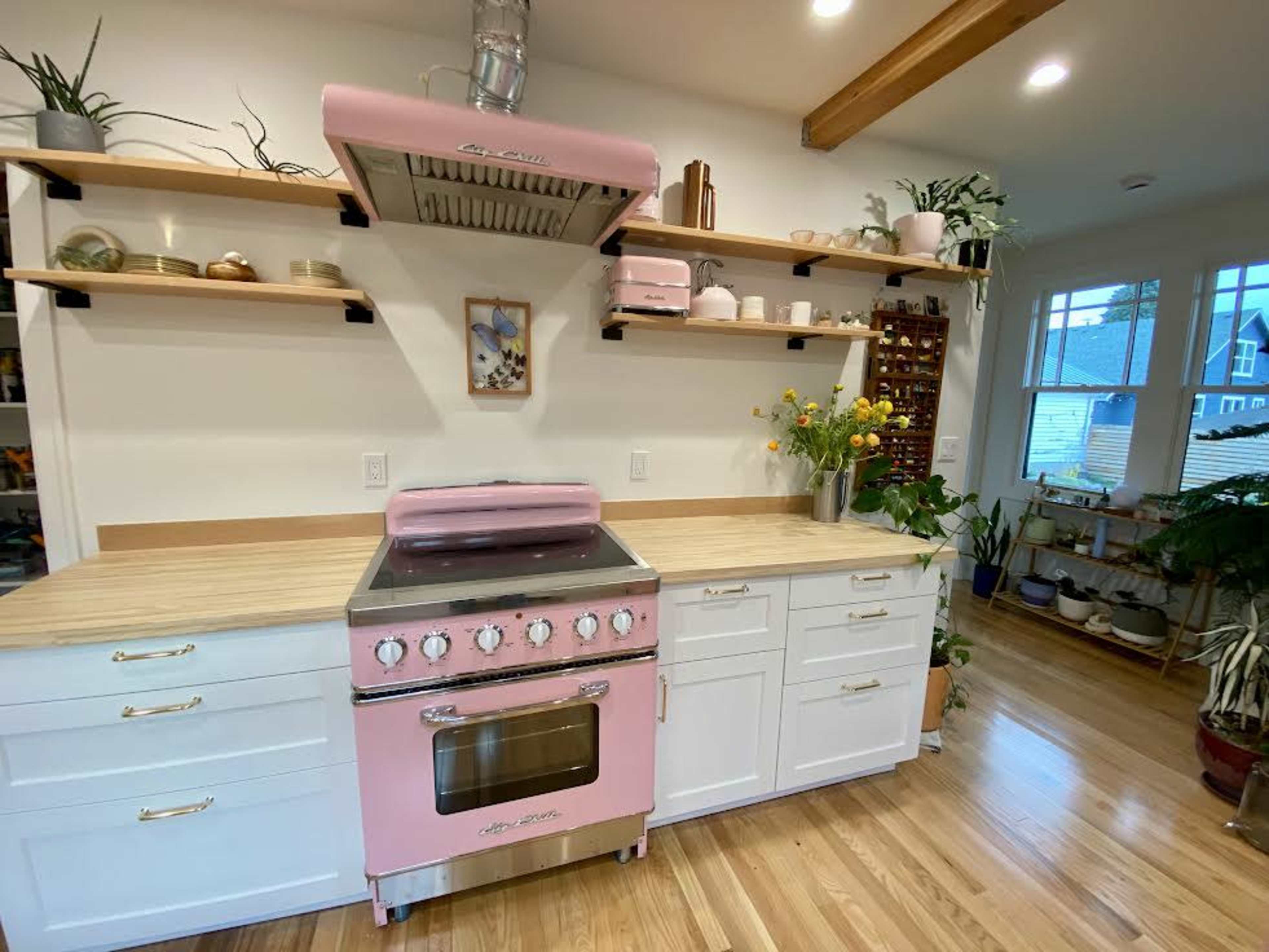 A pink stove is positioned under a hood in a kitchen with wooden shelves displaying plants and kitchenware against white cabinetry.