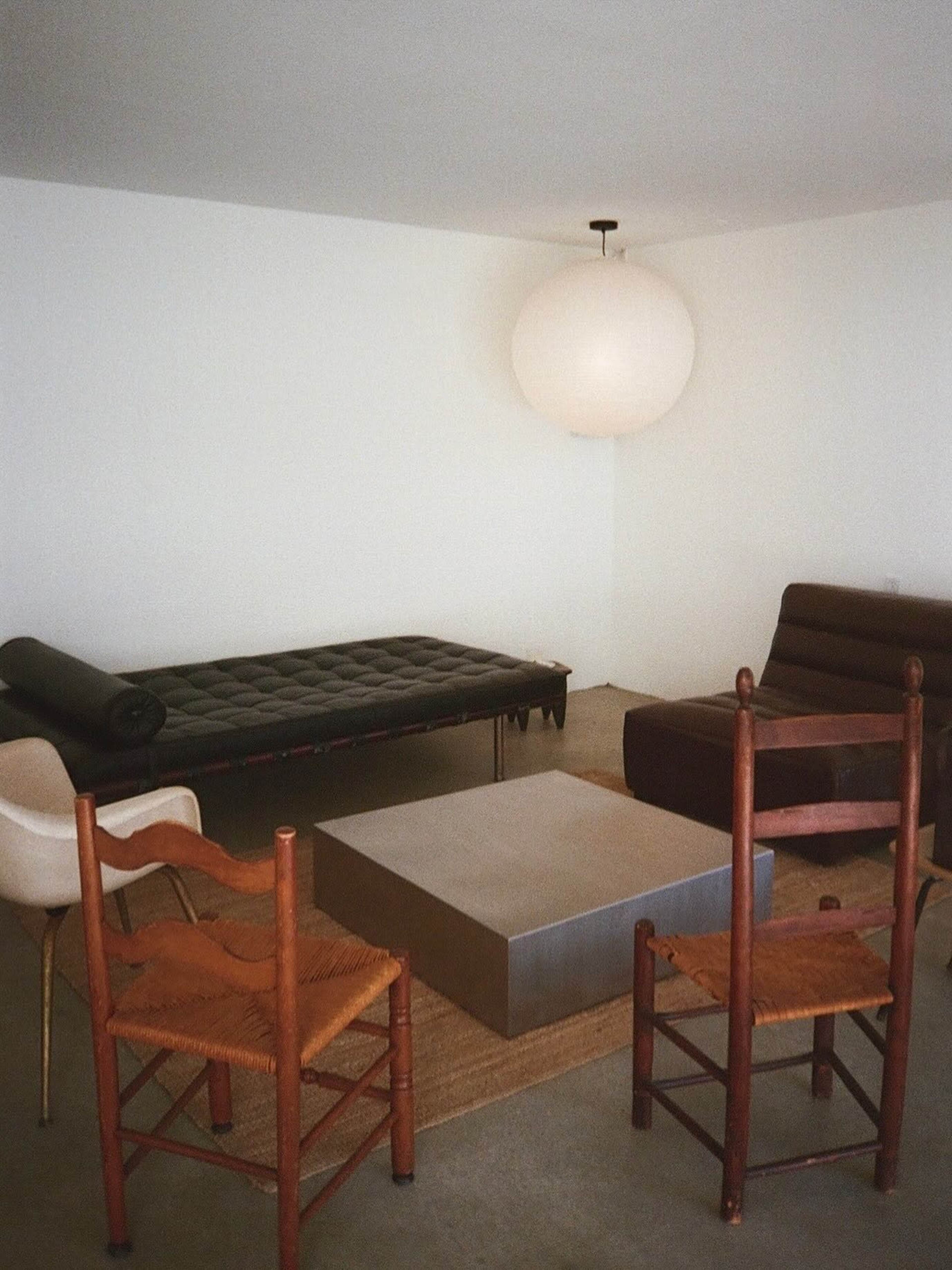 The image shows a minimalist living room featuring a low coffee table surrounded by a mix of four chairs, including two wooden ones and one streamlined, modern chair, under a large pendant light.