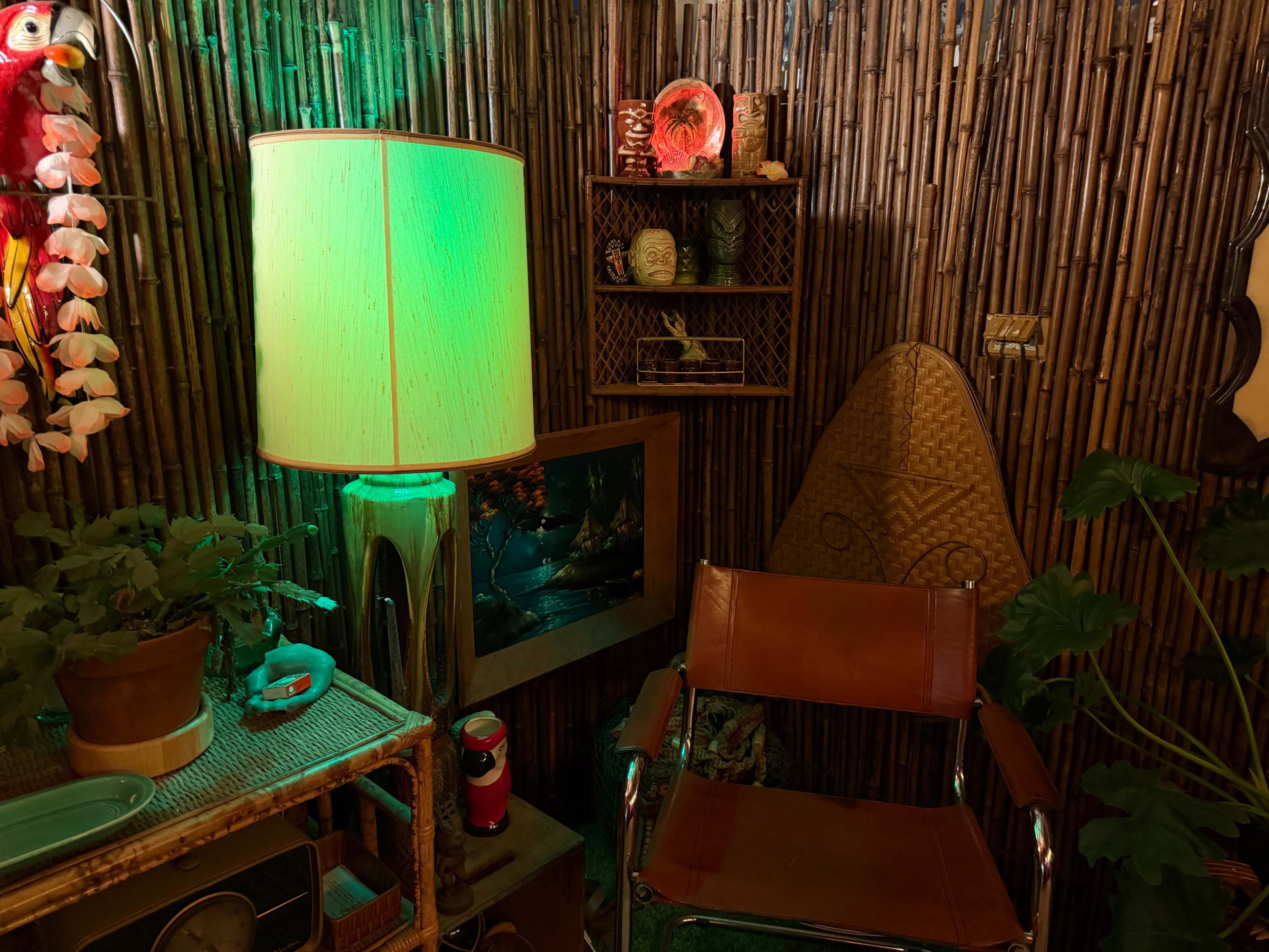 The image shows a cozy corner of a room with bamboo walls, a green-lit lamp, a wooden chair, and various decorative items including plants and artwork.
