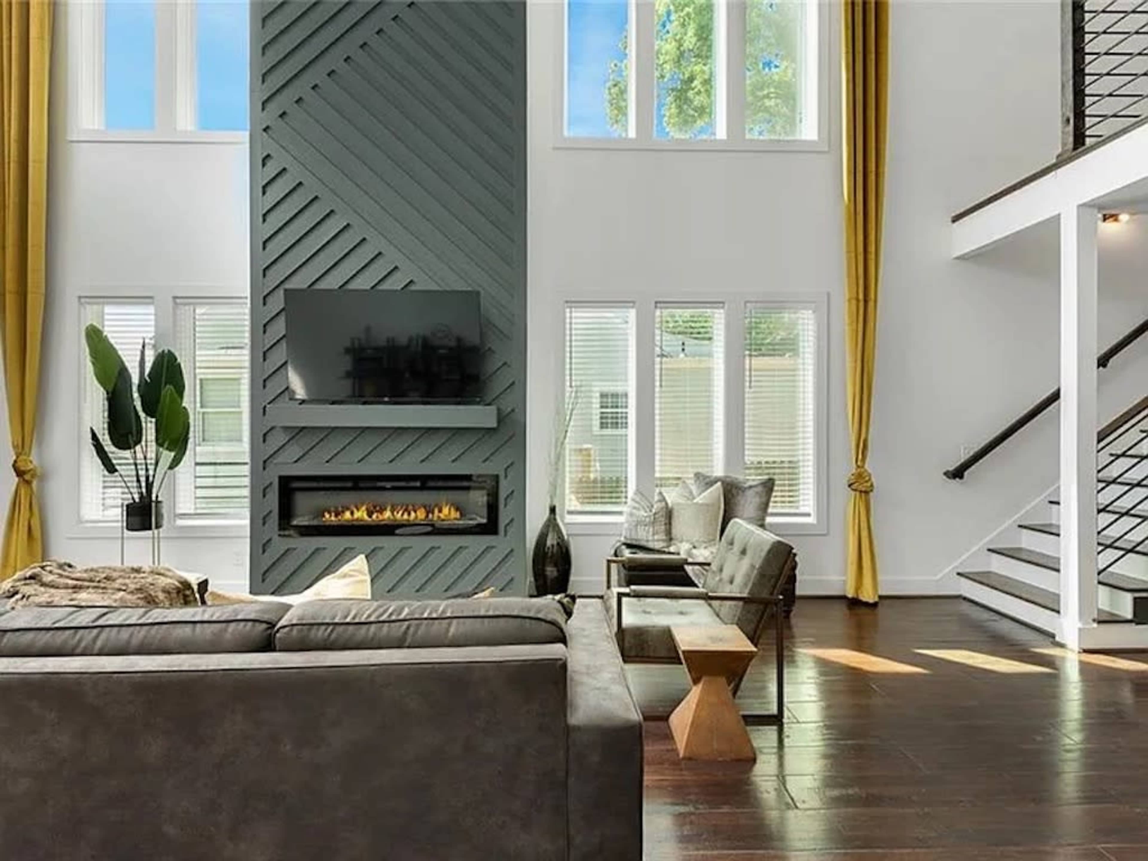 A modern living room features a tall fireplace with a TV mounted above it, surrounded by large windows and yellow curtains, alongside a sofa and minimalist furniture.