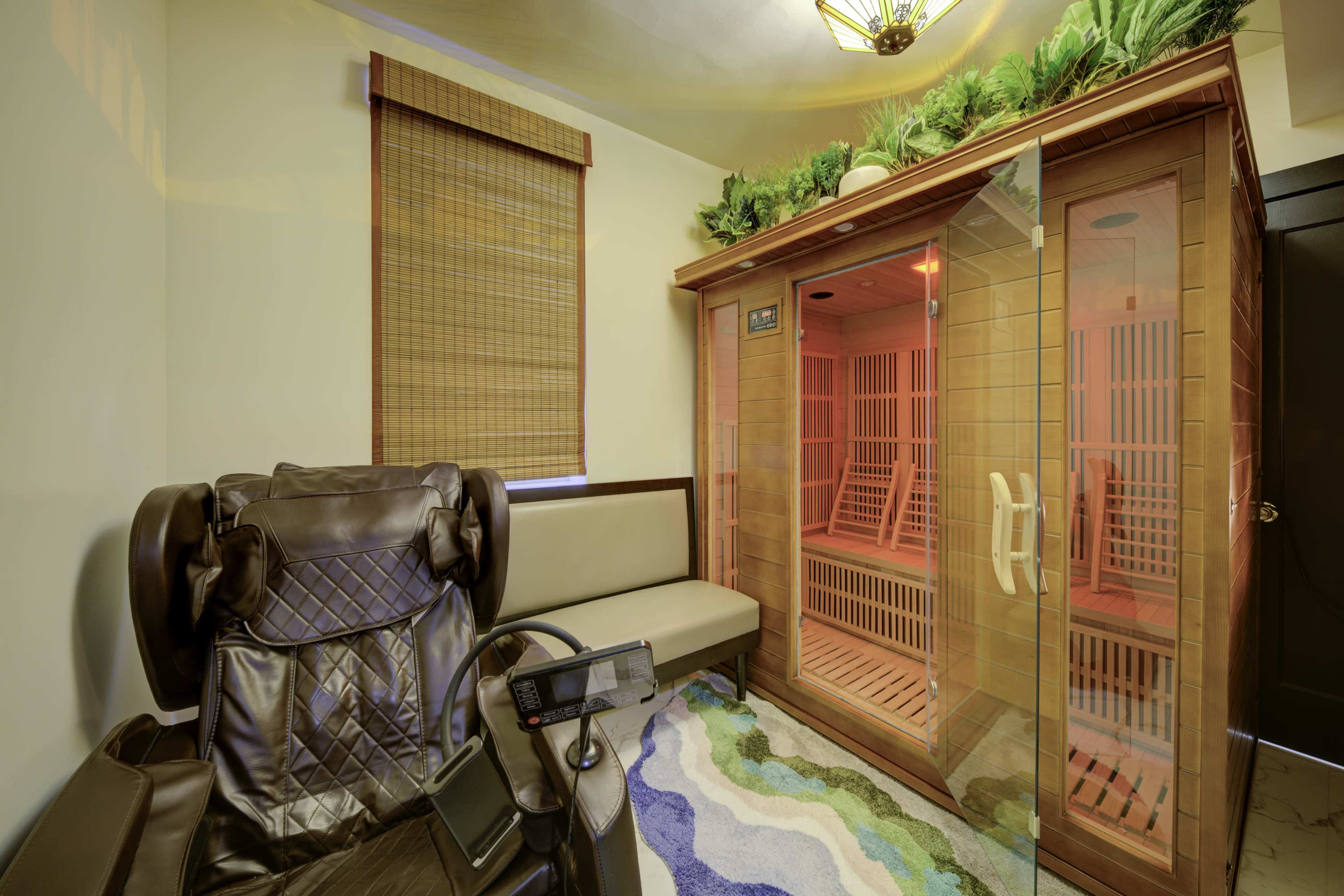 The image features a corner of a wellness room containing a glass infrared sauna and a massage chair, with natural light filtering through a window covered by bamboo blinds.
