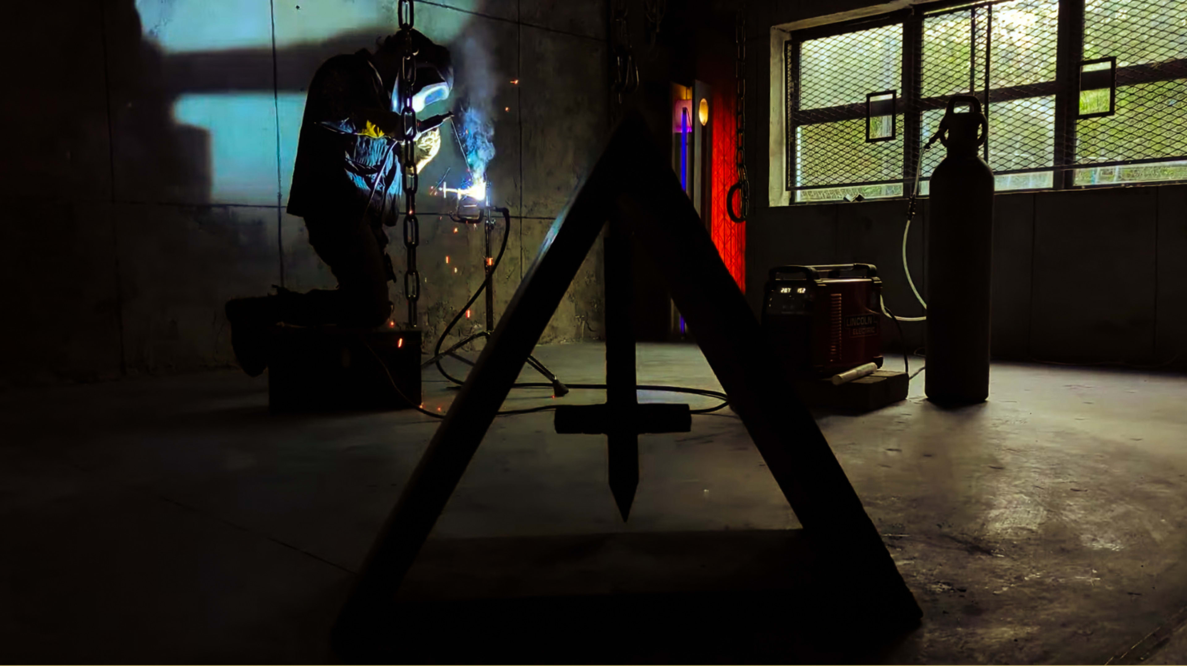 A welder, with a protective helmet, is kneeling and working on a metal piece in a dimly lit industrial space, while sparks fly from the welding process.