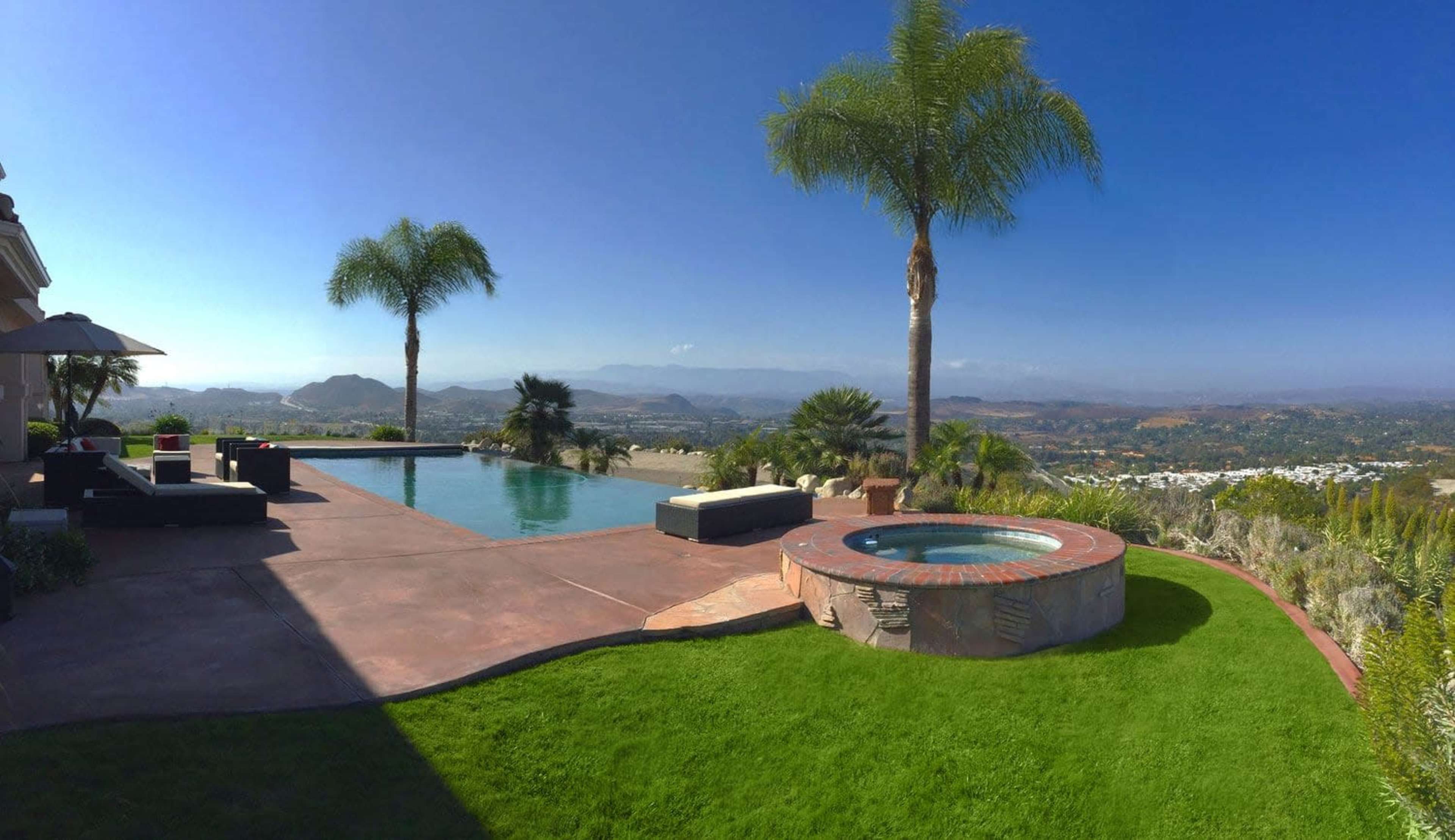 The image shows a modern outdoor space with a swimming pool, spa, manicured lawn, and palm trees, overlooking a scenic view of hills and valleys.