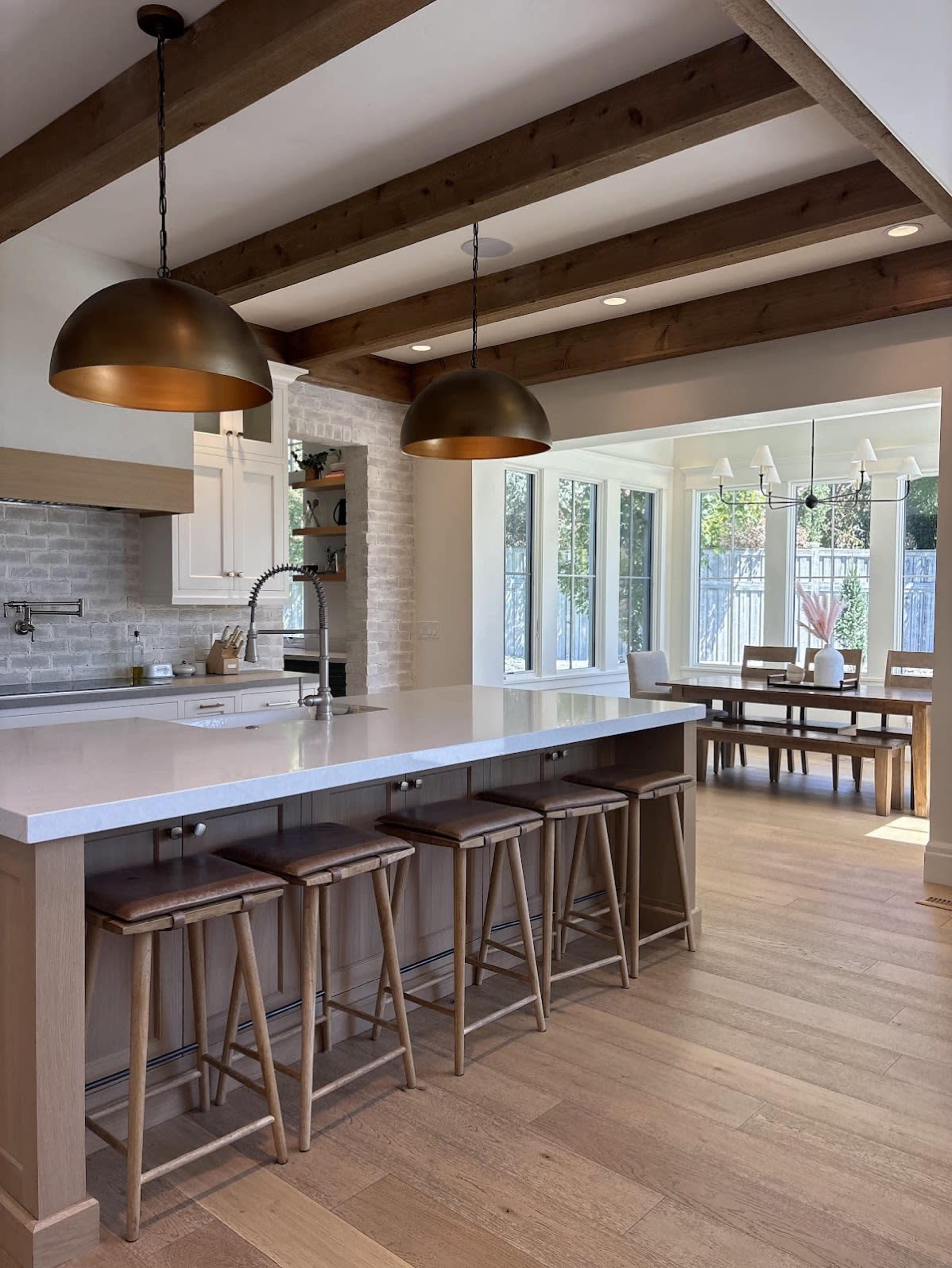 A modern kitchen features an island with wooden stools, pendant lighting, and large windows overlooking a dining area.