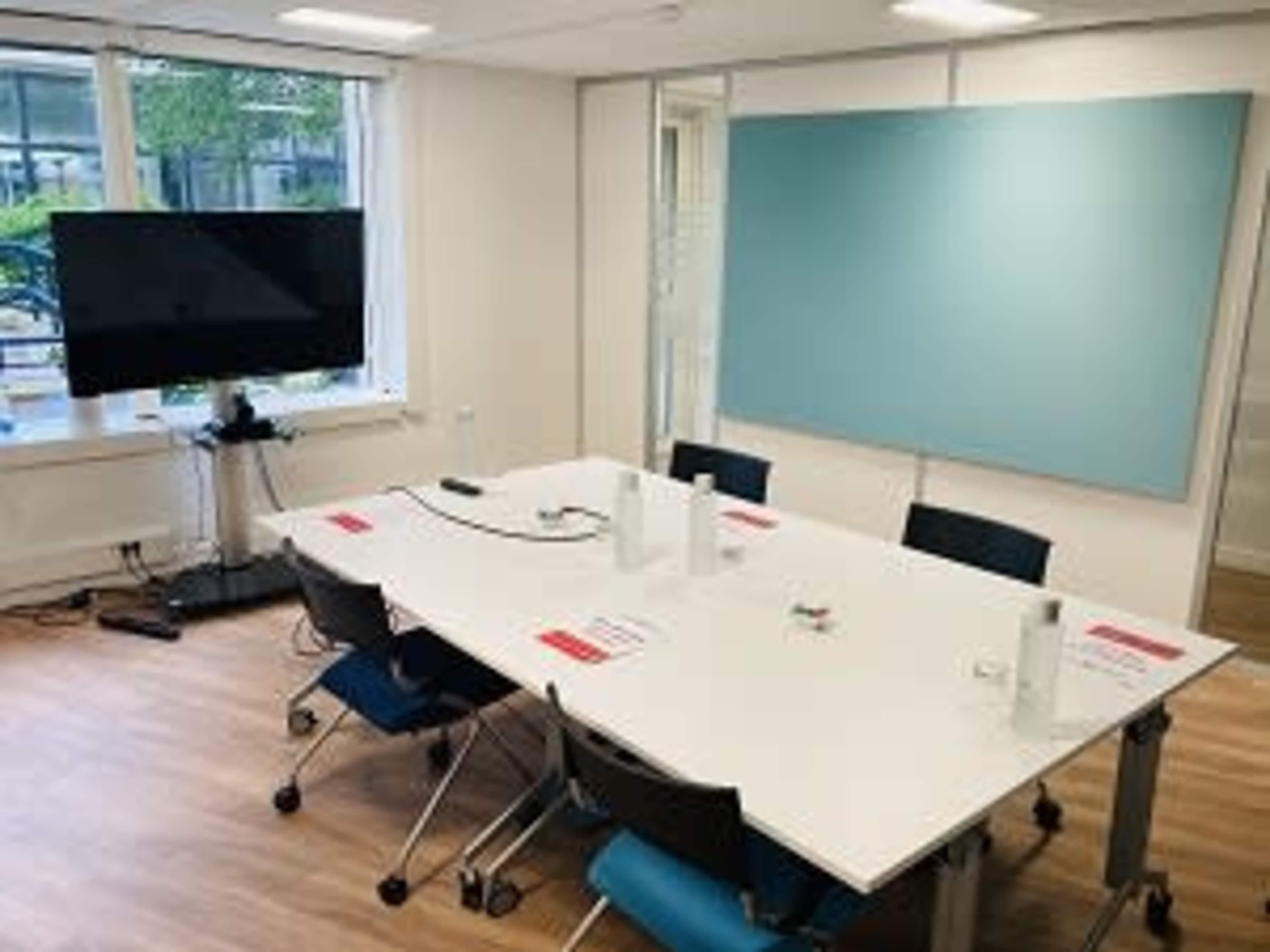 A meeting room features a large white table surrounded by chairs, equipped with a television on a stand and a blue wall area for presentations.