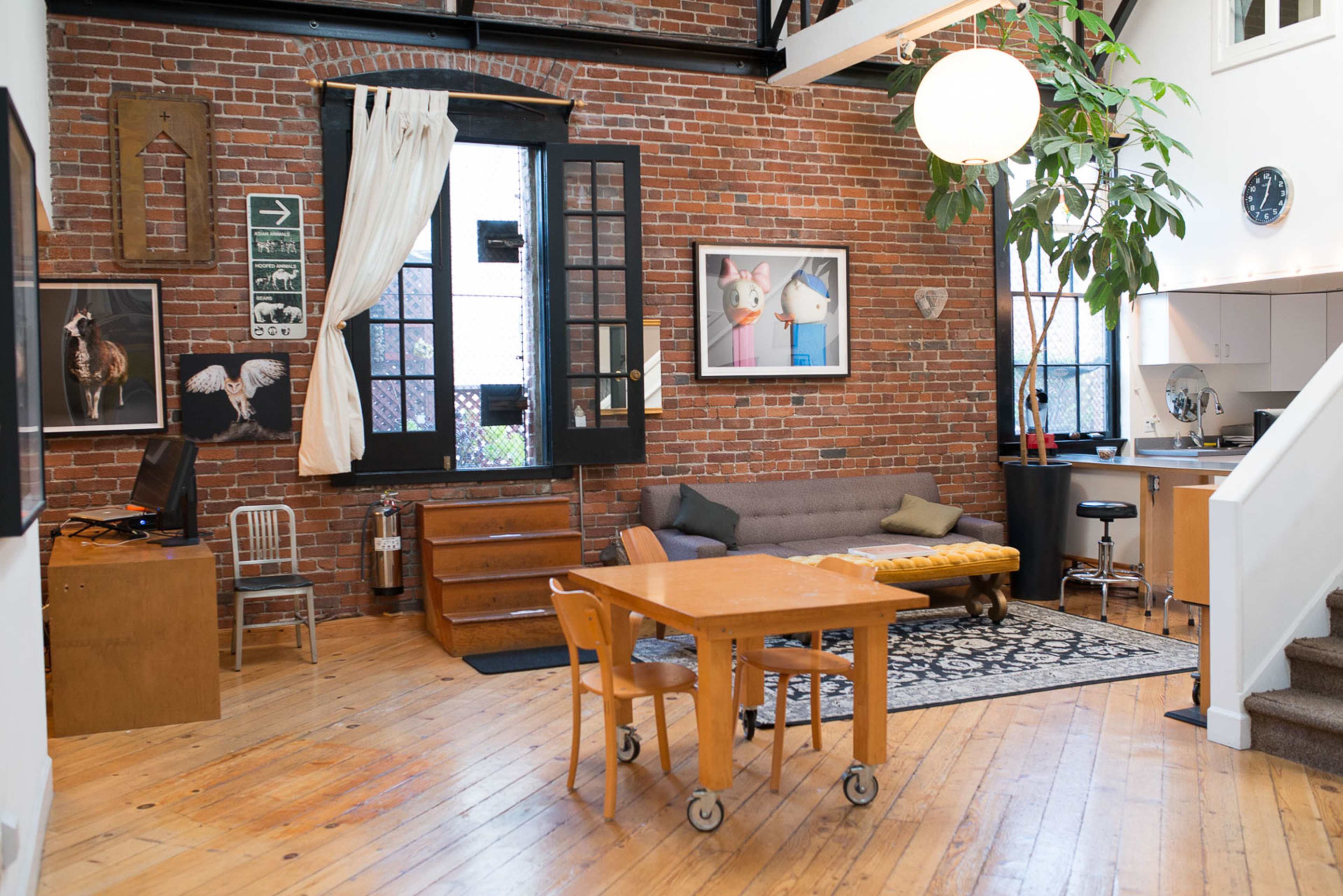 The image shows a modern loft-style living space with exposed brick walls, a large window, a gray sofa, a wooden dining table, and a small kitchen area.