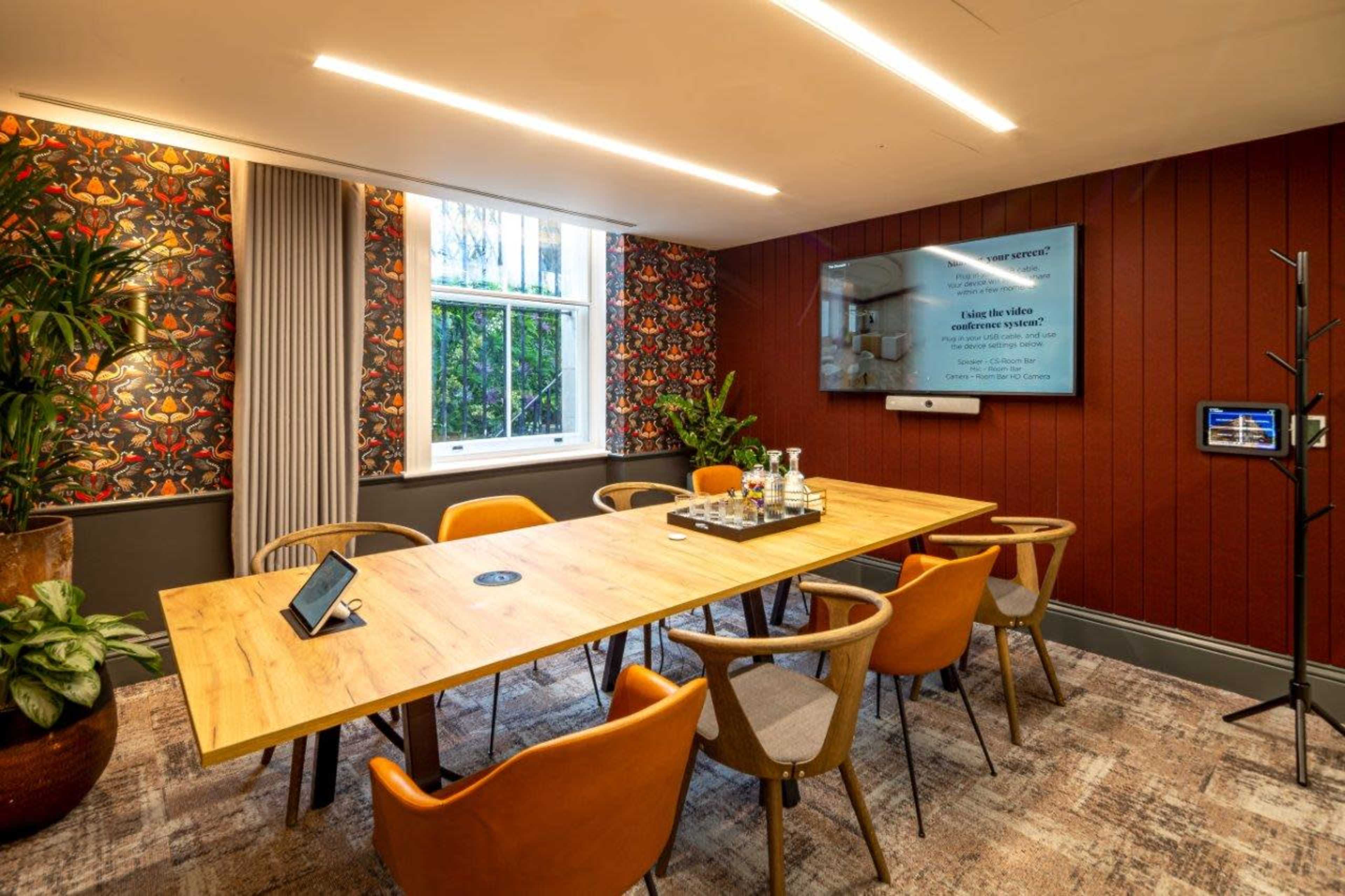 A modern meeting room featuring a long wooden table surrounded by six chairs, potted plants, a large screen on the wall, and decorative wallpaper.