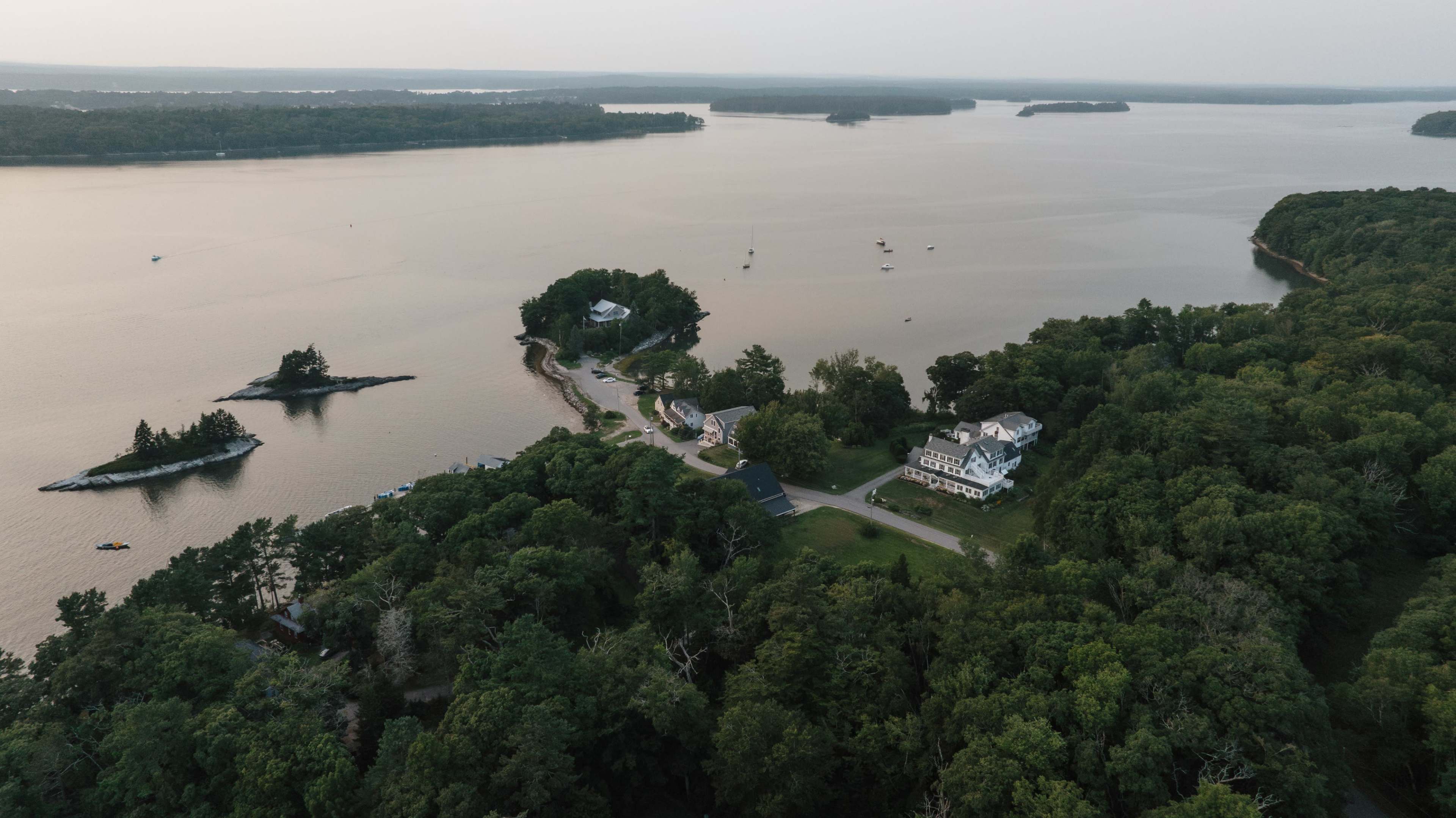 The image captures an aerial view of a coastal landscape featuring a cluster of homes along the shoreline, surrounded by dense greenery and a calm water body.