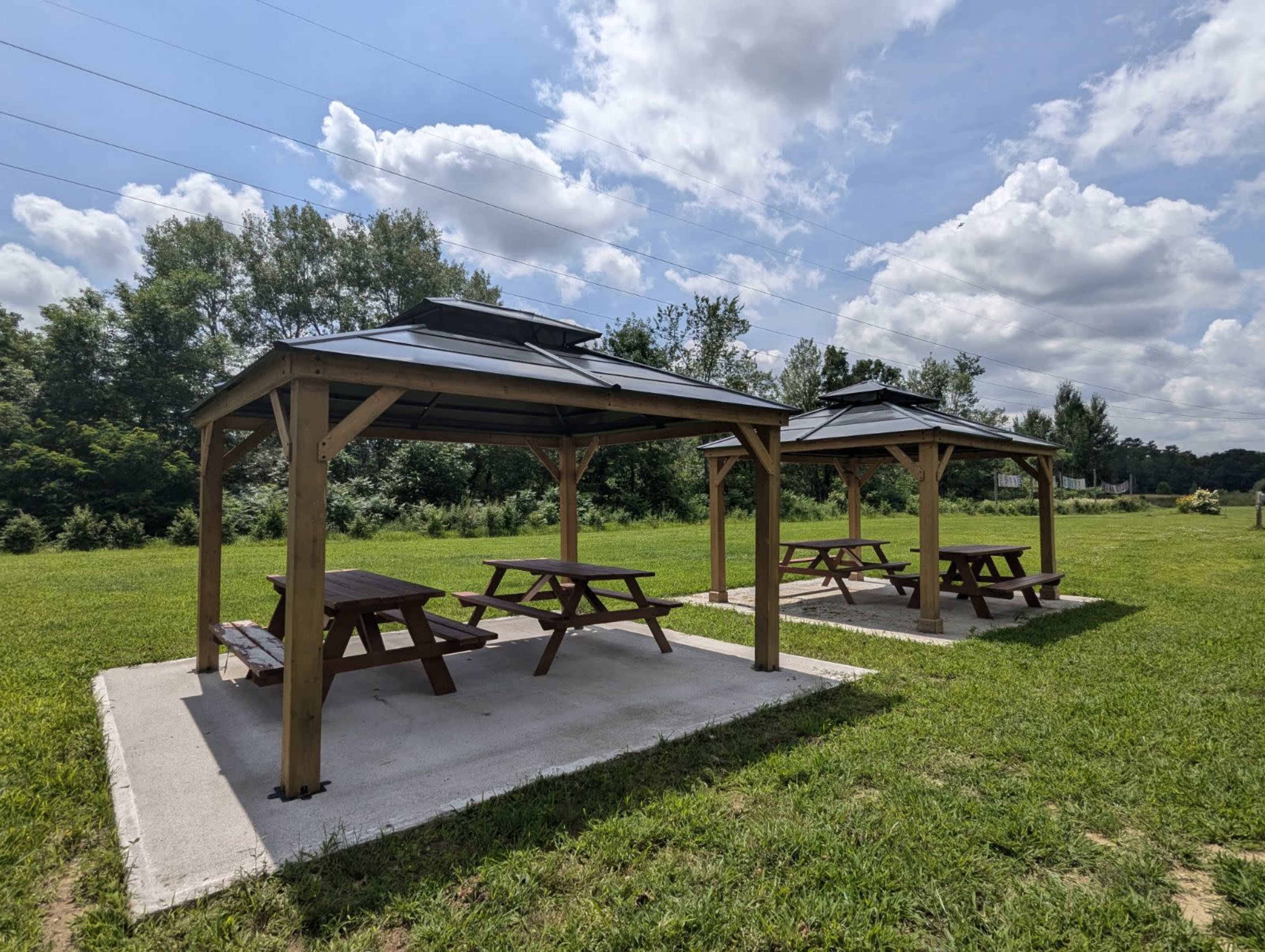 The image shows two wooden gazebos with picnic tables on a grassy area, surrounded by trees and under a partly cloudy sky.