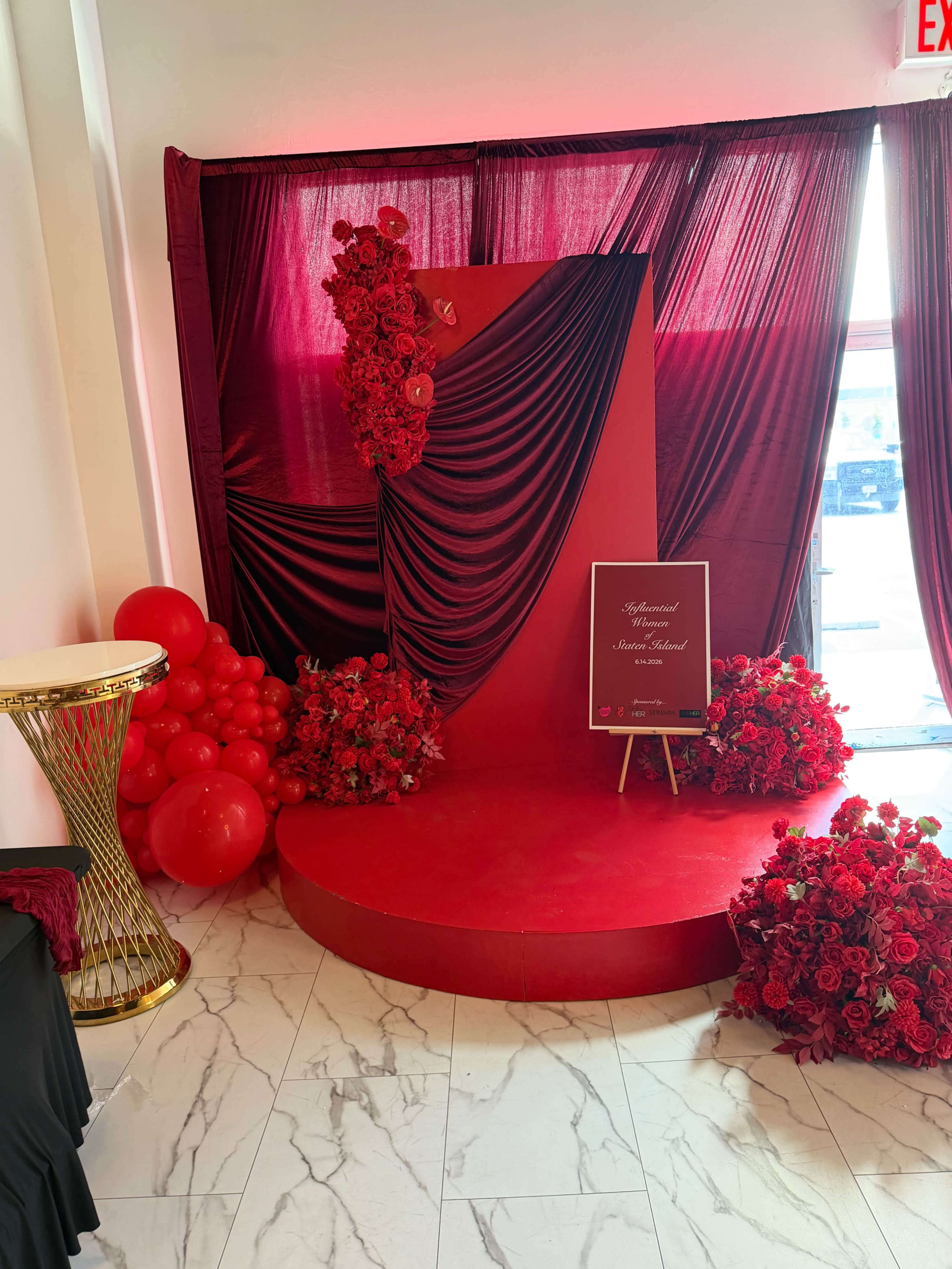 The image depicts a red-themed event backdrop featuring draped fabric, floral arrangements, and a circular platform, set against a marble floor.