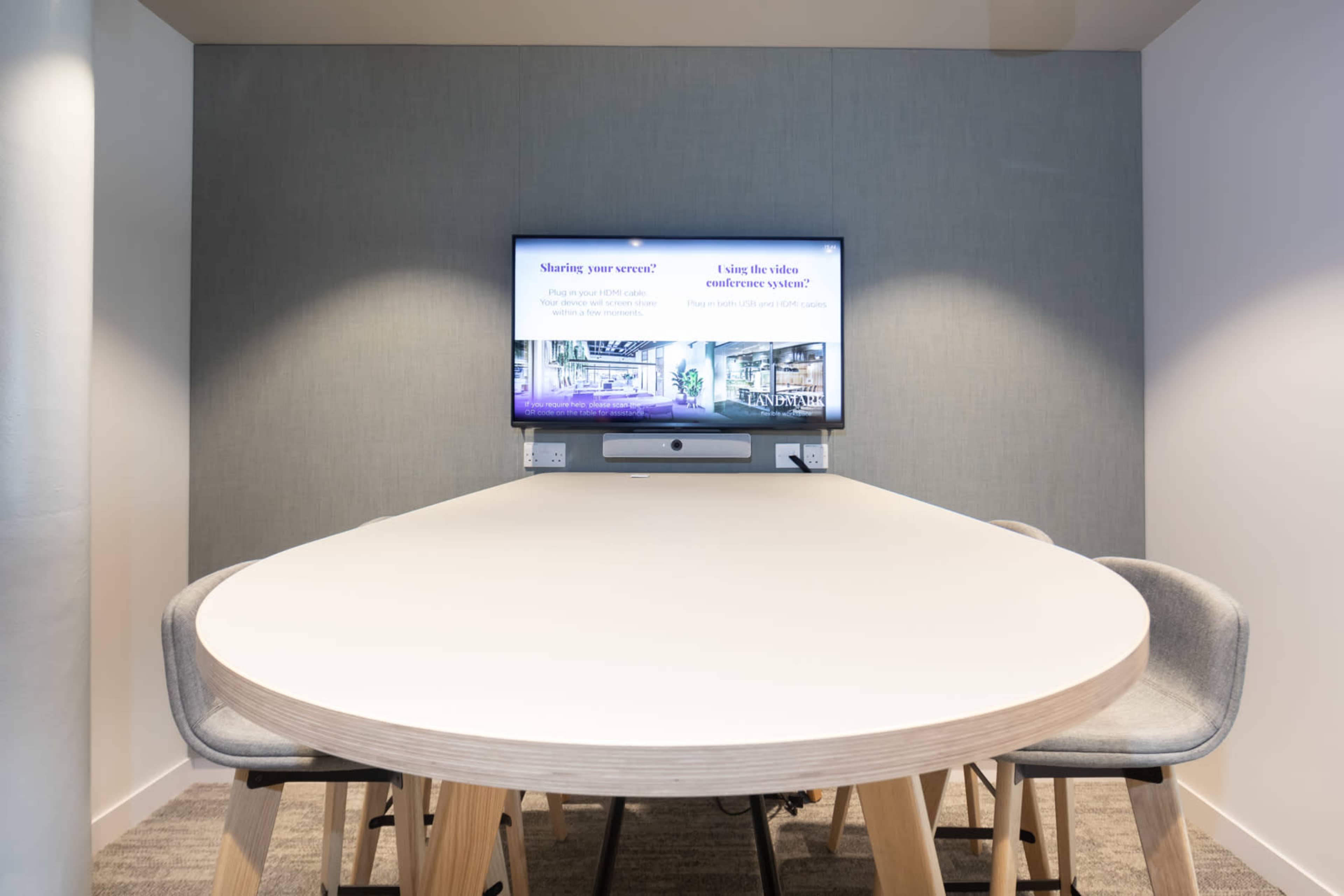 A modern conference room features a large oval table surrounded by gray chairs, with a screen displaying a presentation on the wall.
