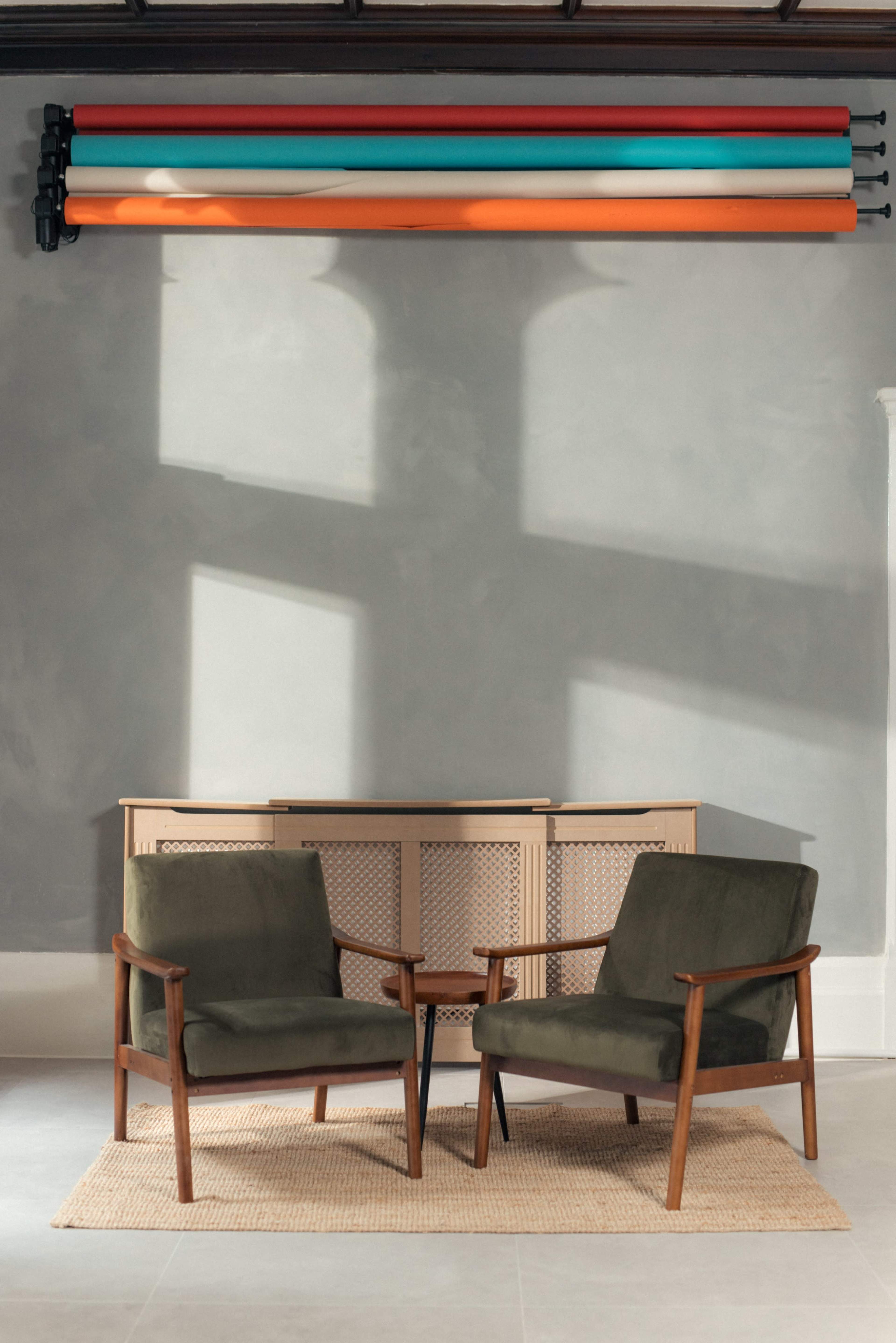 Two green armchairs are positioned across from each other on a light floor, with a cabinet behind them and colorful rolls of material mounted on the wall above.