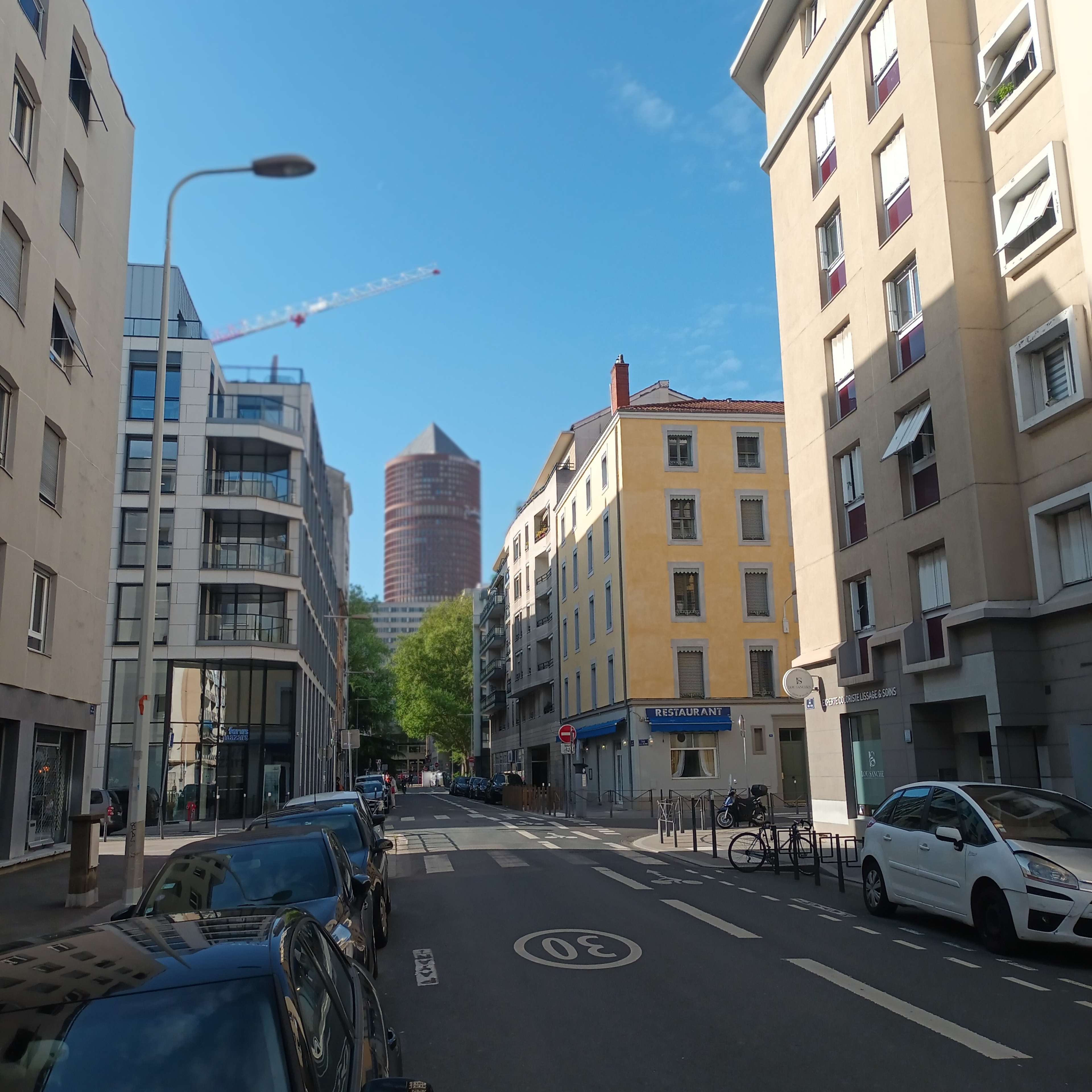 The image shows a street lined with residential buildings and parked cars, with a tower visible in the background under a clear blue sky.