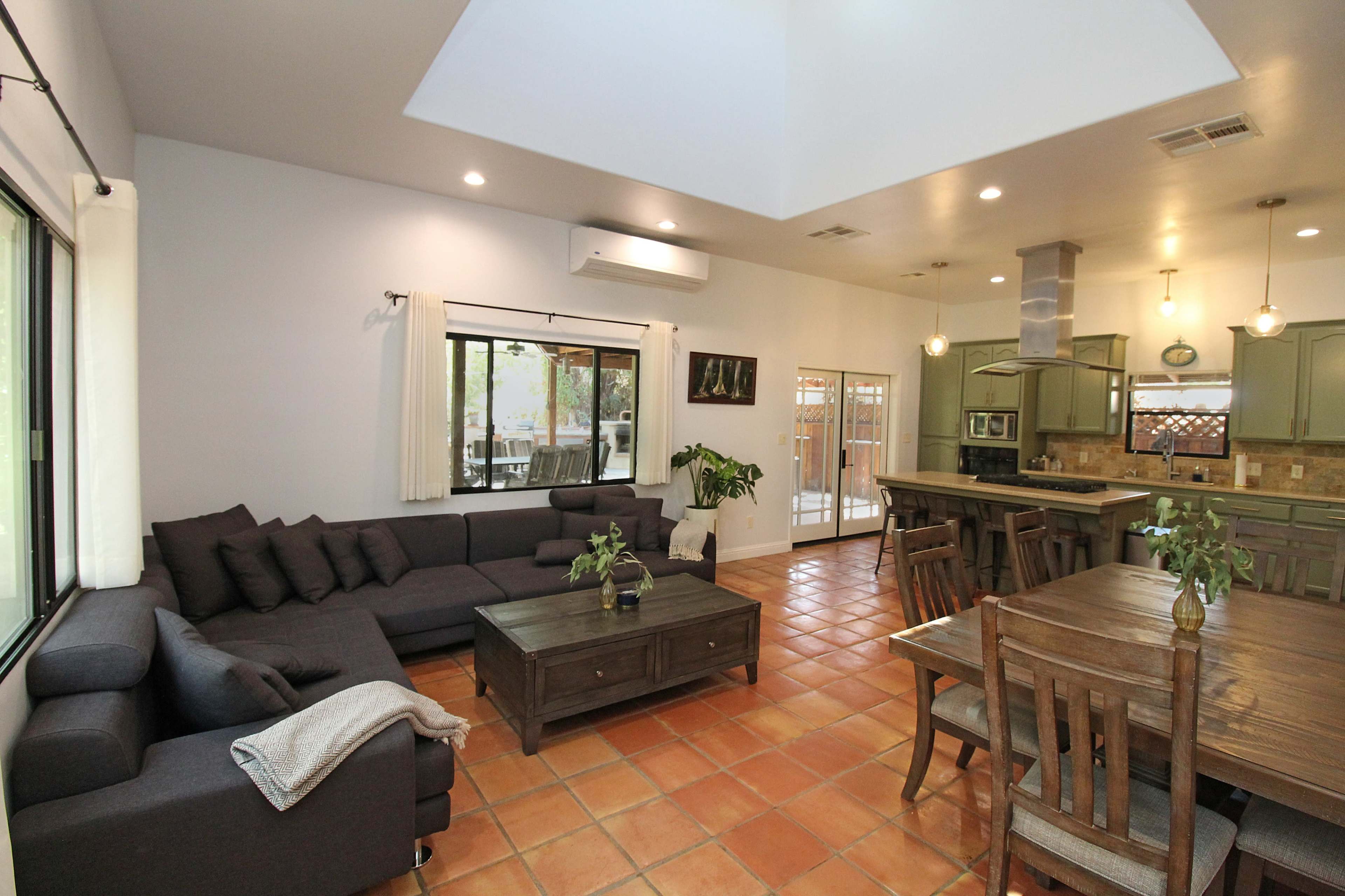 A spacious living area with a dark sectional sofa, a wooden coffee table, and a dining table, all featuring terracotta tile flooring.