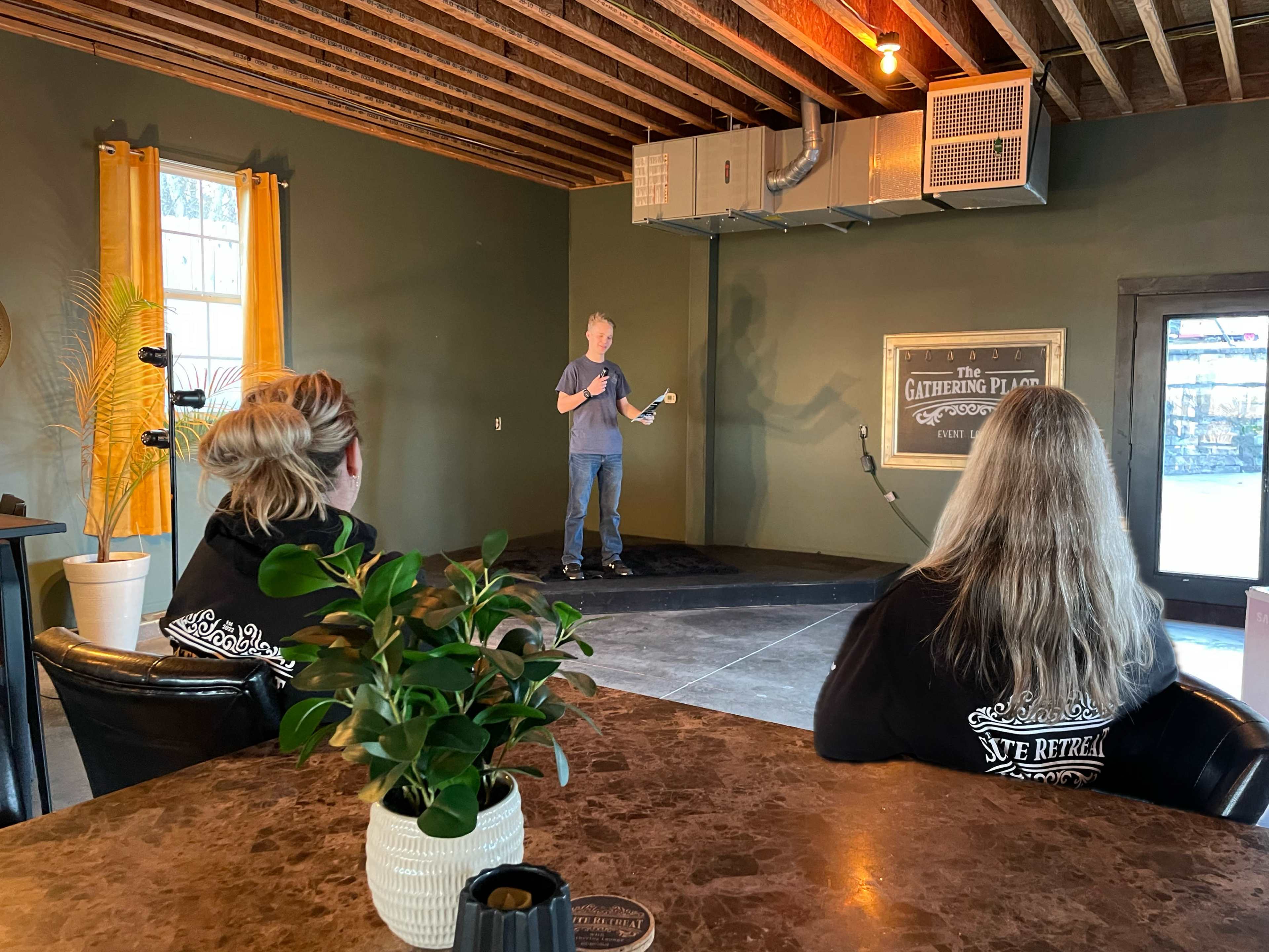 A speaker stands on a small stage in a venue while two people sit at a table nearby, listening attentively.