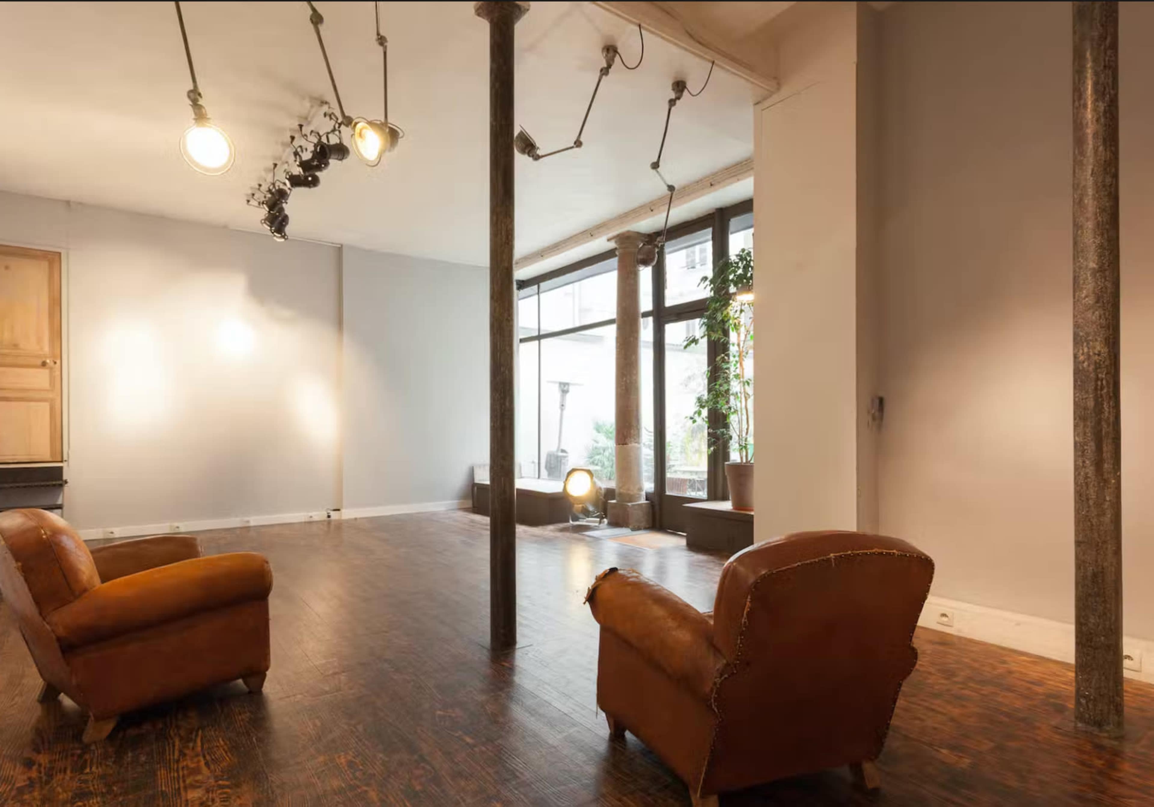 The image shows a minimalist interior space with two leather armchairs facing each other, wooden flooring, and large windows letting in natural light.