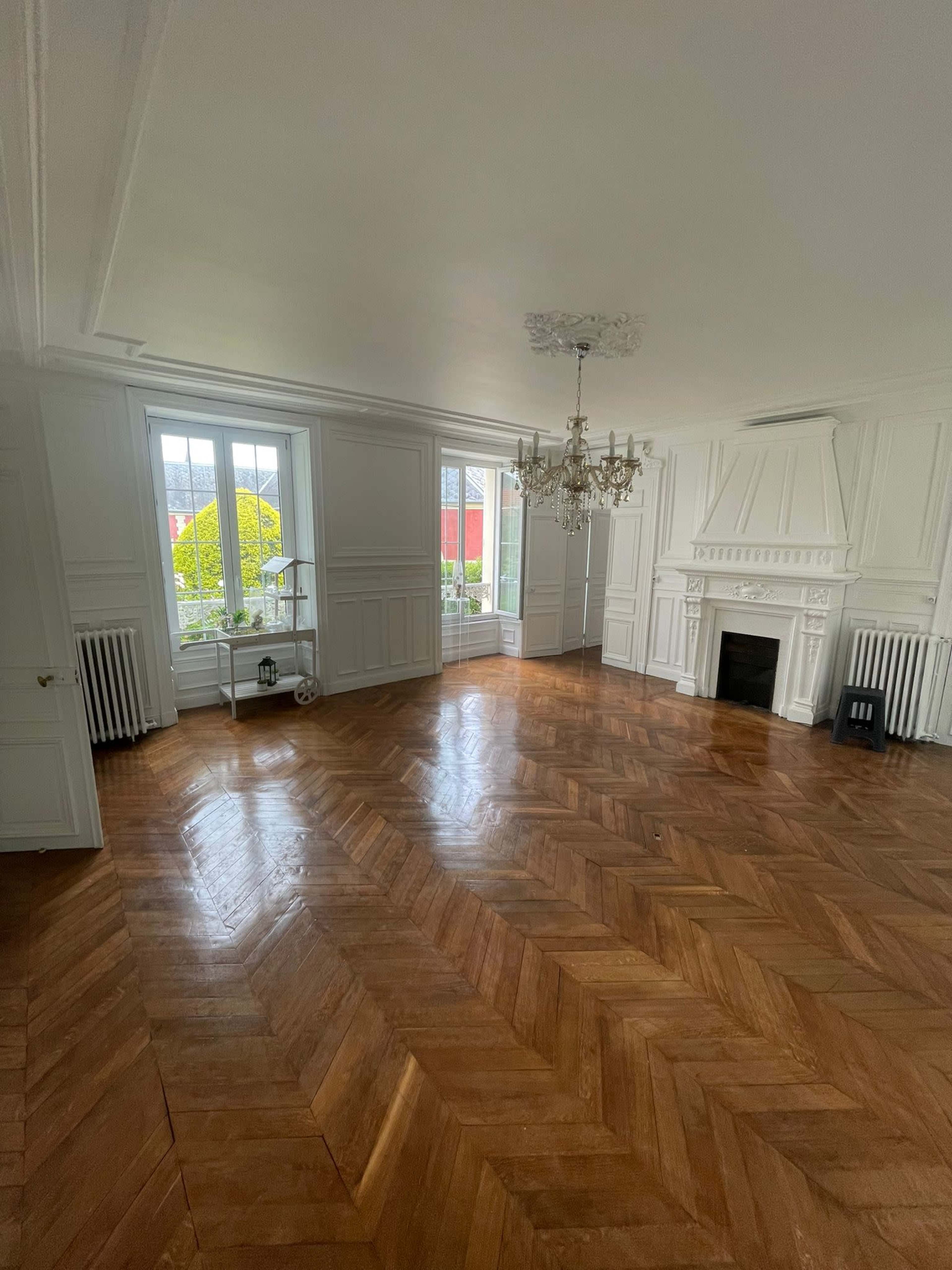 A well-lit, empty room with herringbone wooden flooring, white paneled walls, and a chandelier hanging from the ceiling.
