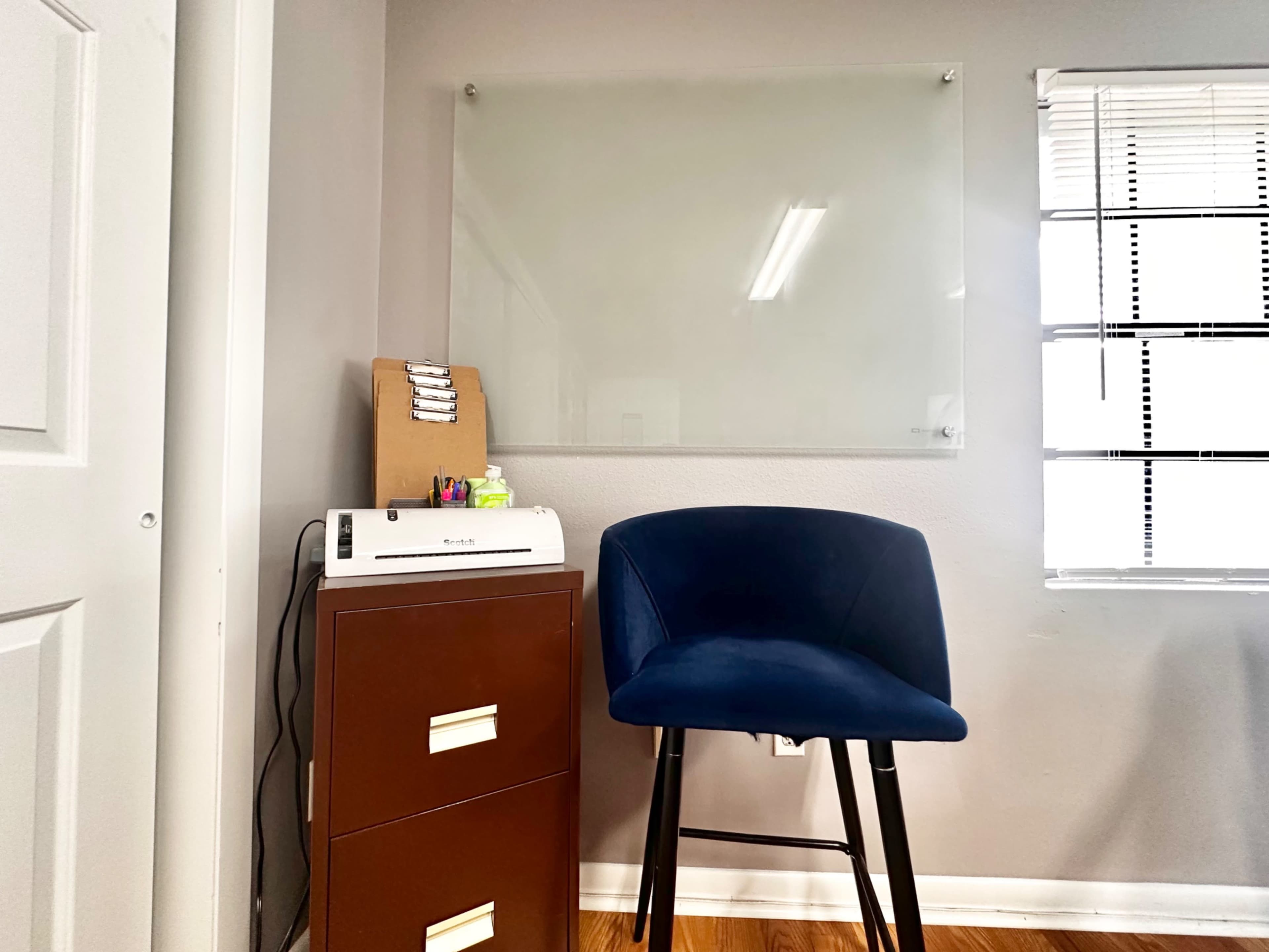 The image shows a small office space featuring a blue chair next to a brown filing cabinet, with a clear whiteboard mounted on the wall and a window with horizontal blinds.