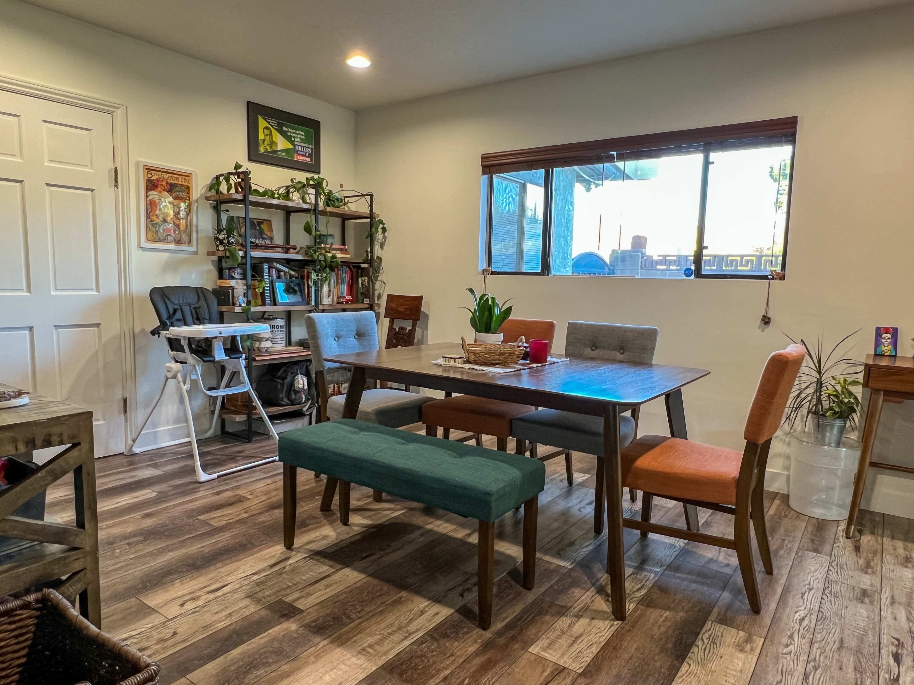 The image shows a dining area with a wooden table surrounded by chairs of various colors, a high chair, and decorative plants against the walls.