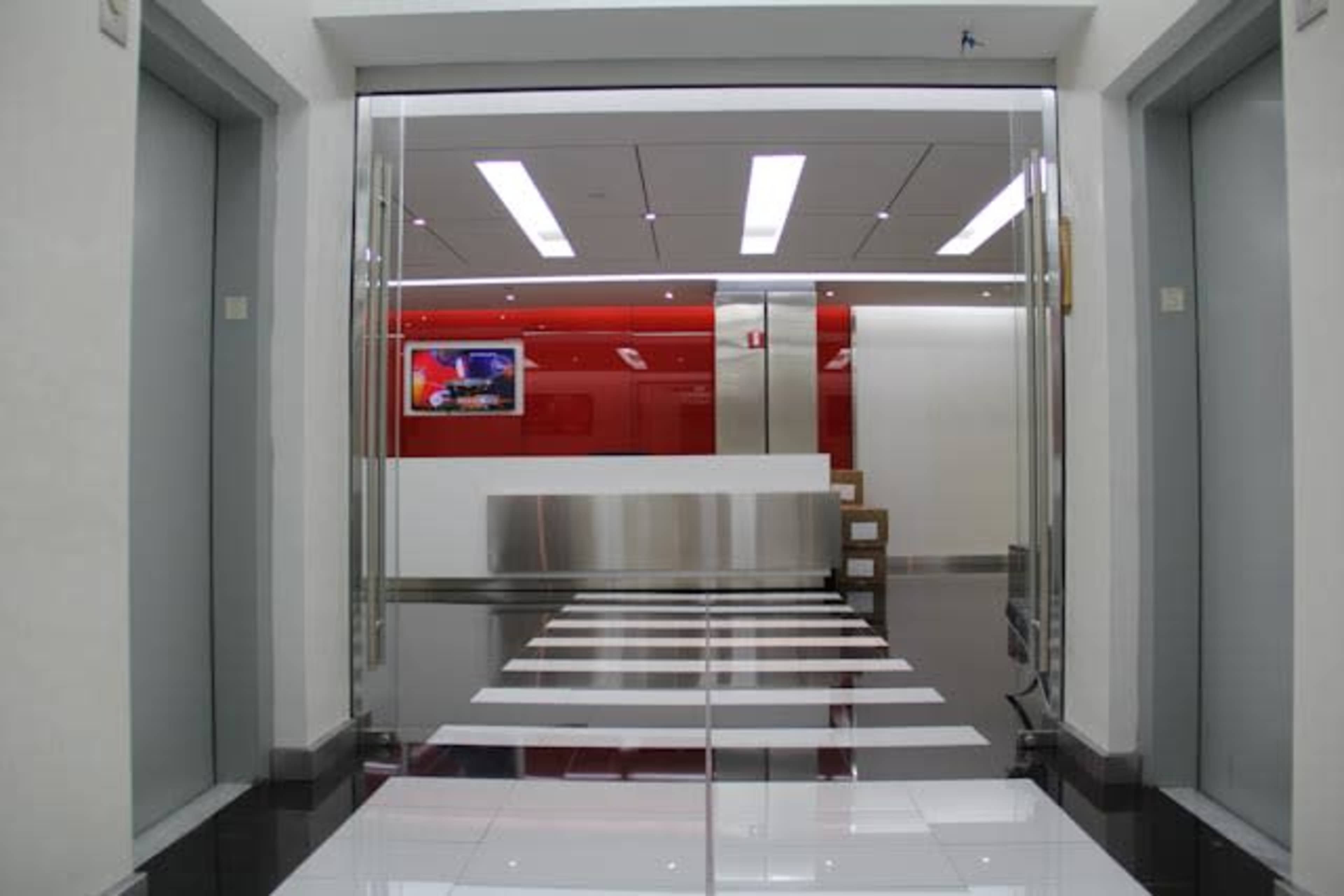 The image shows a modern lobby area with glass doors leading to a reception desk, flanked by two elevator doors.