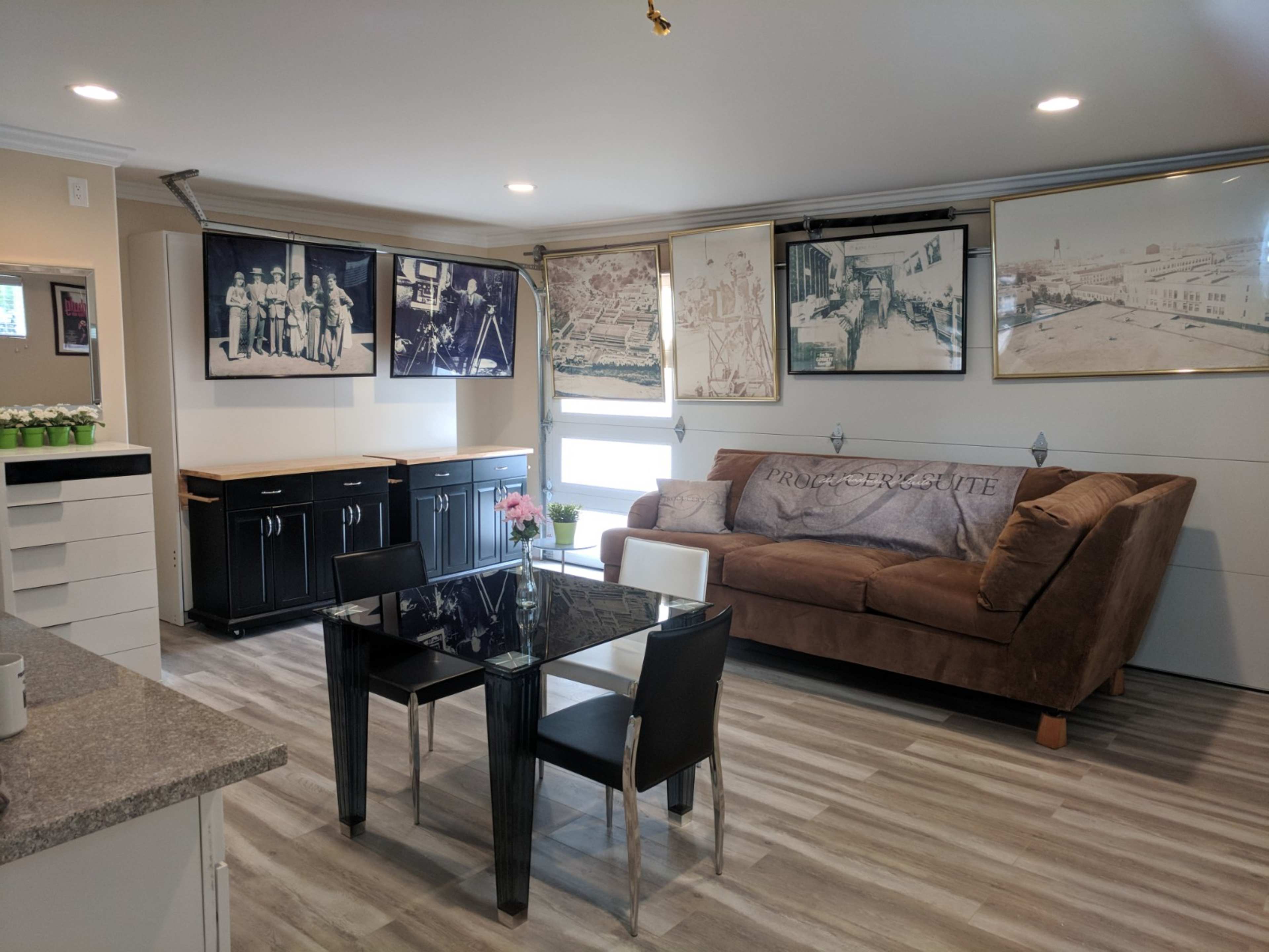 The image shows a living area with a brown sofa, a glass dining table surrounded by black chairs, and framed black-and-white photographs on the walls.
