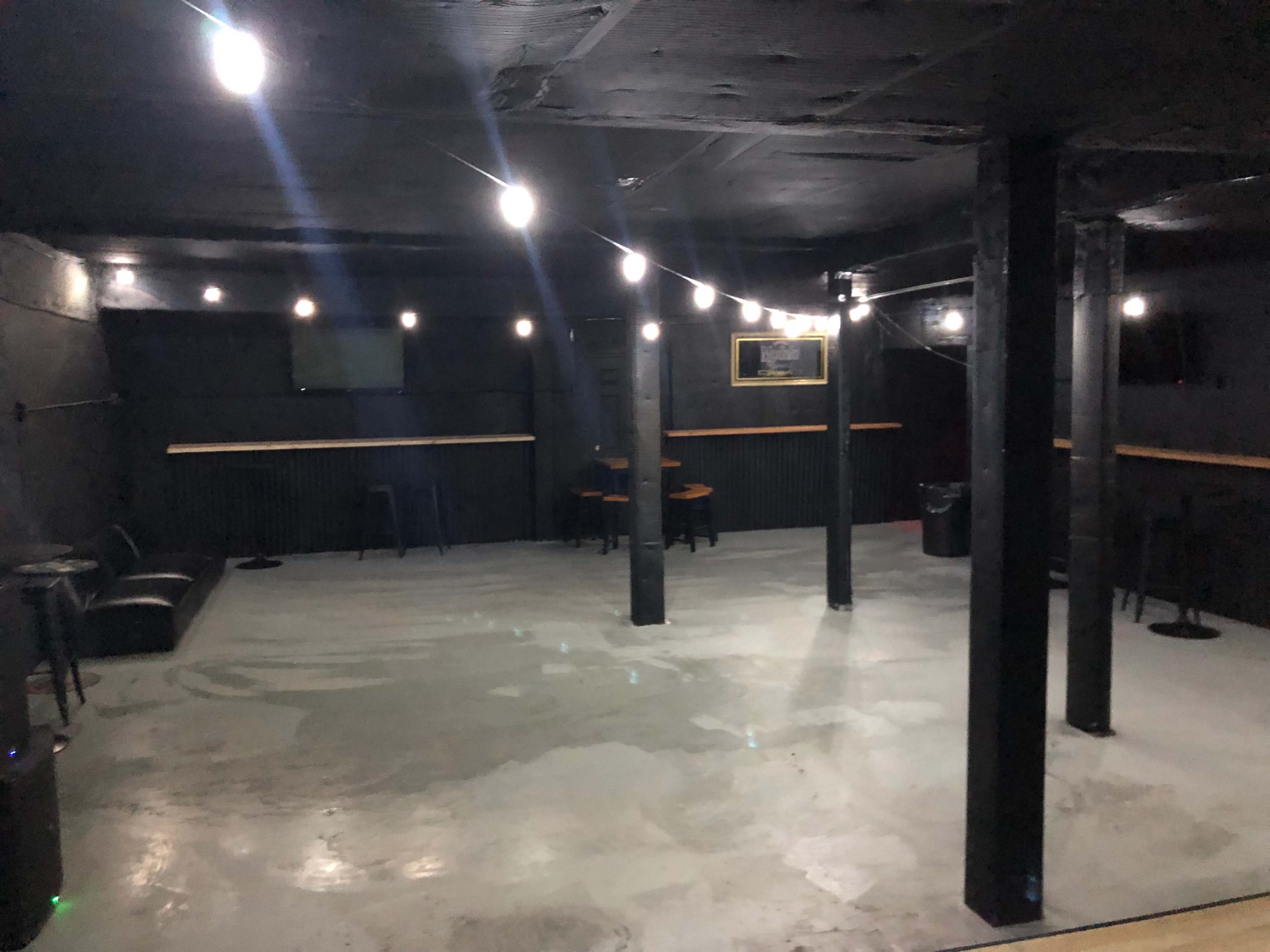 The image shows a dimly lit, sparsely furnished indoor venue with concrete flooring and overhead string lights.