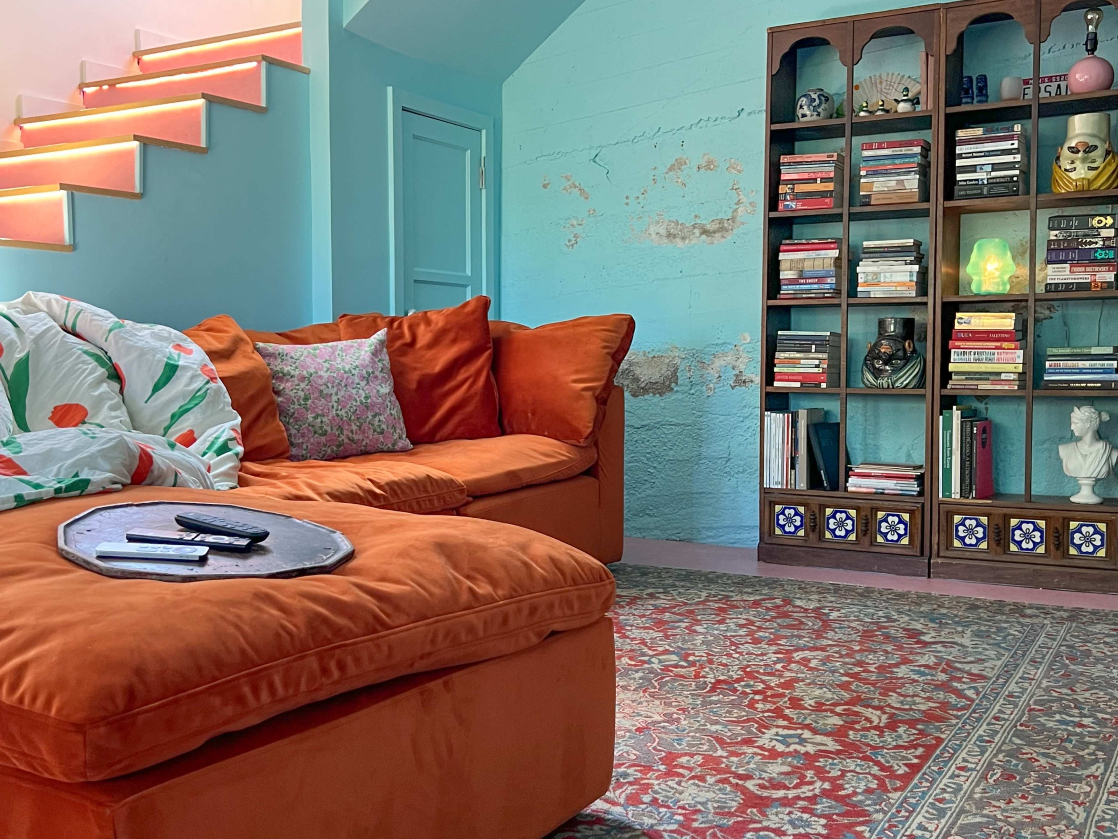 The image shows a living room with an orange sofa, a colorful rug, a bookshelf filled with books and decorative items, and a staircase in the background.