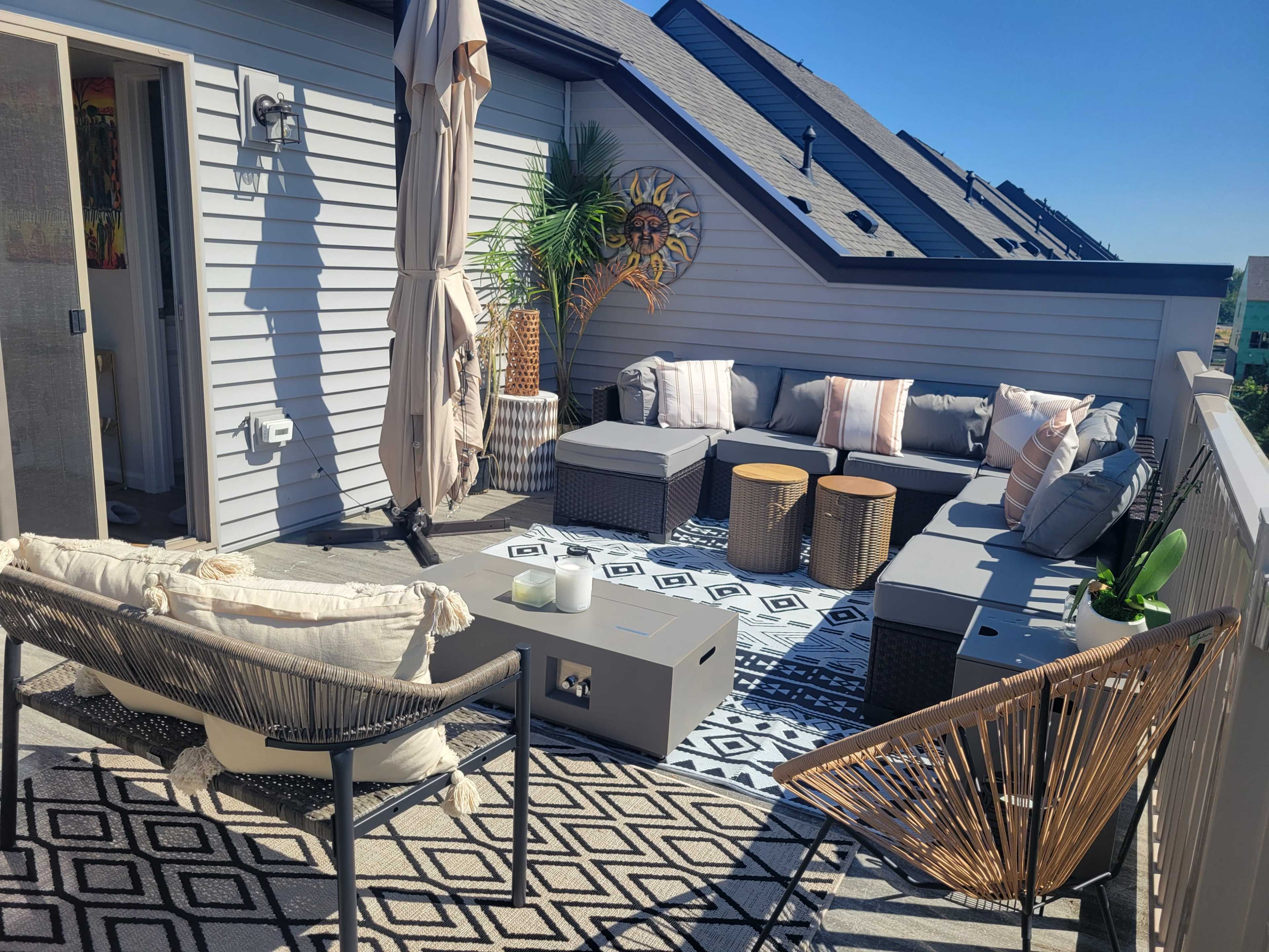 The image shows a rooftop patio area featuring outdoor seating with cushions, a patterned rug, decorative plants, and sunlight illuminating the space.