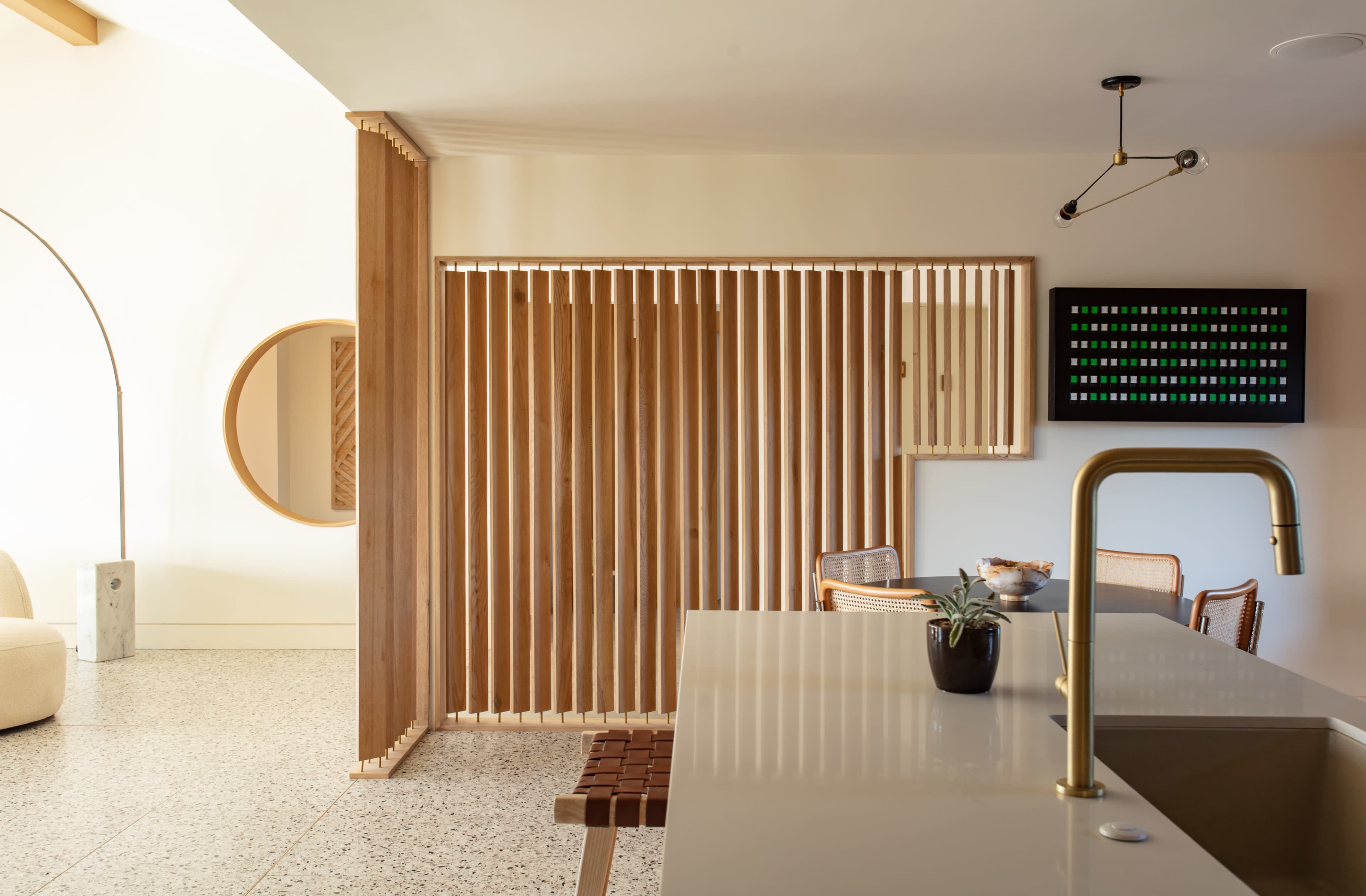 A modern interior space with wooden slat partitions separating a dining area from a kitchen, alongside minimalist decor and a large round mirror.