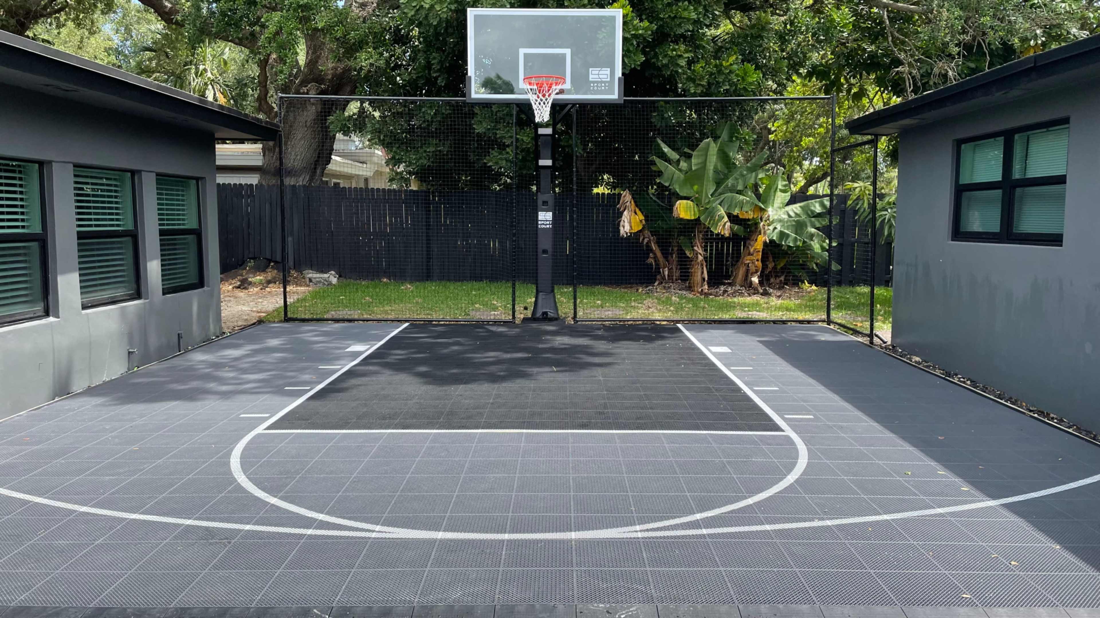 The image shows a basketball court with a hoop and net surrounded by greenery and two buildings.