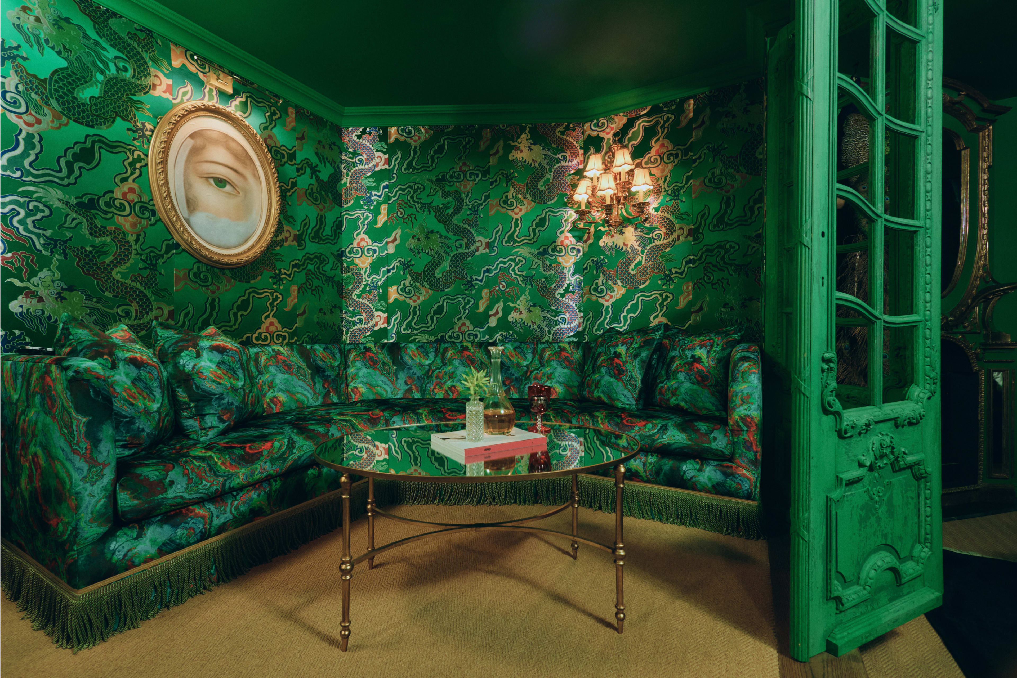 The image shows a vibrant green lounge area with an ornate round couch, a glass table, and a painting on the wall.